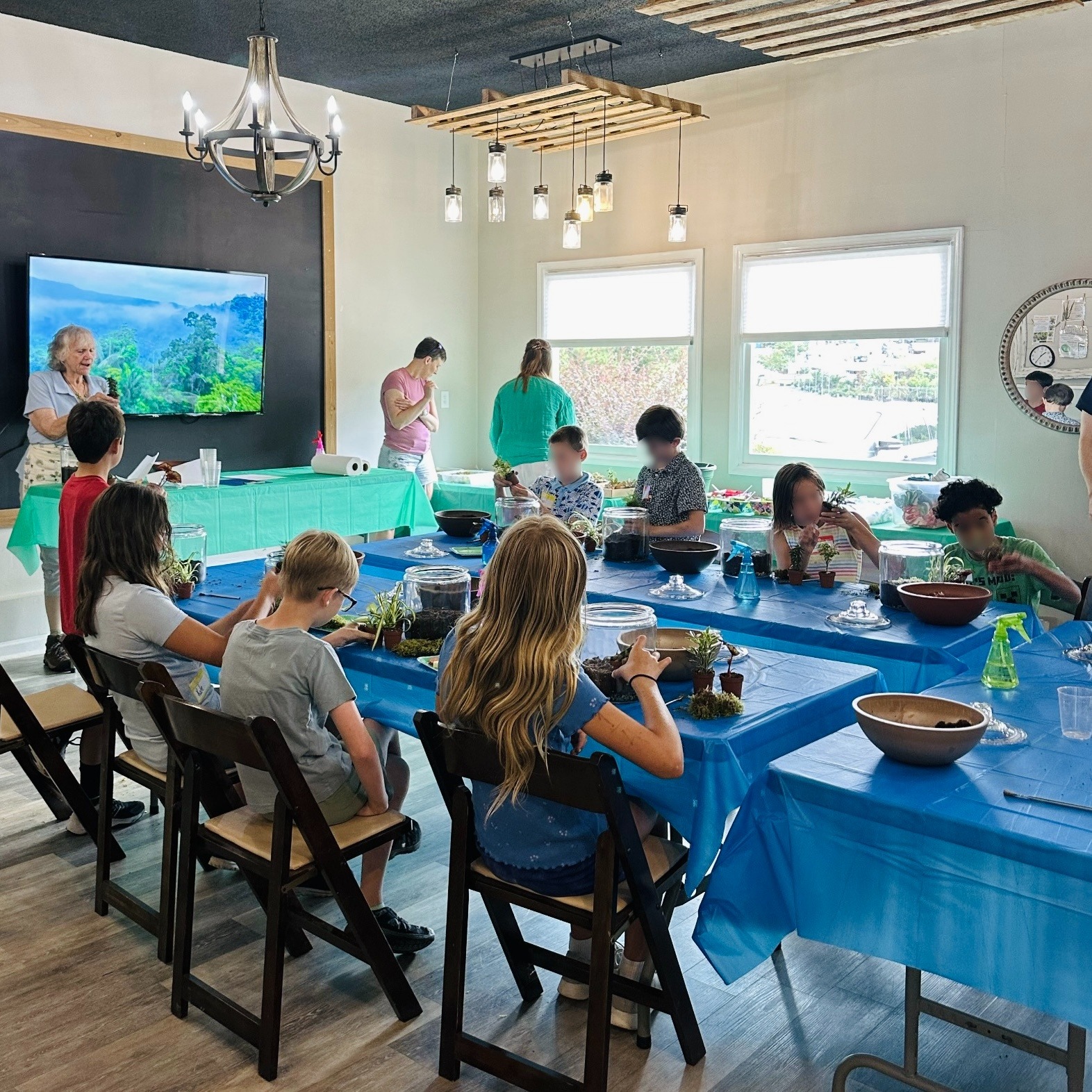 A group of children and adults engage in a crafting activity around a table in a well-lit room with blue table covers.