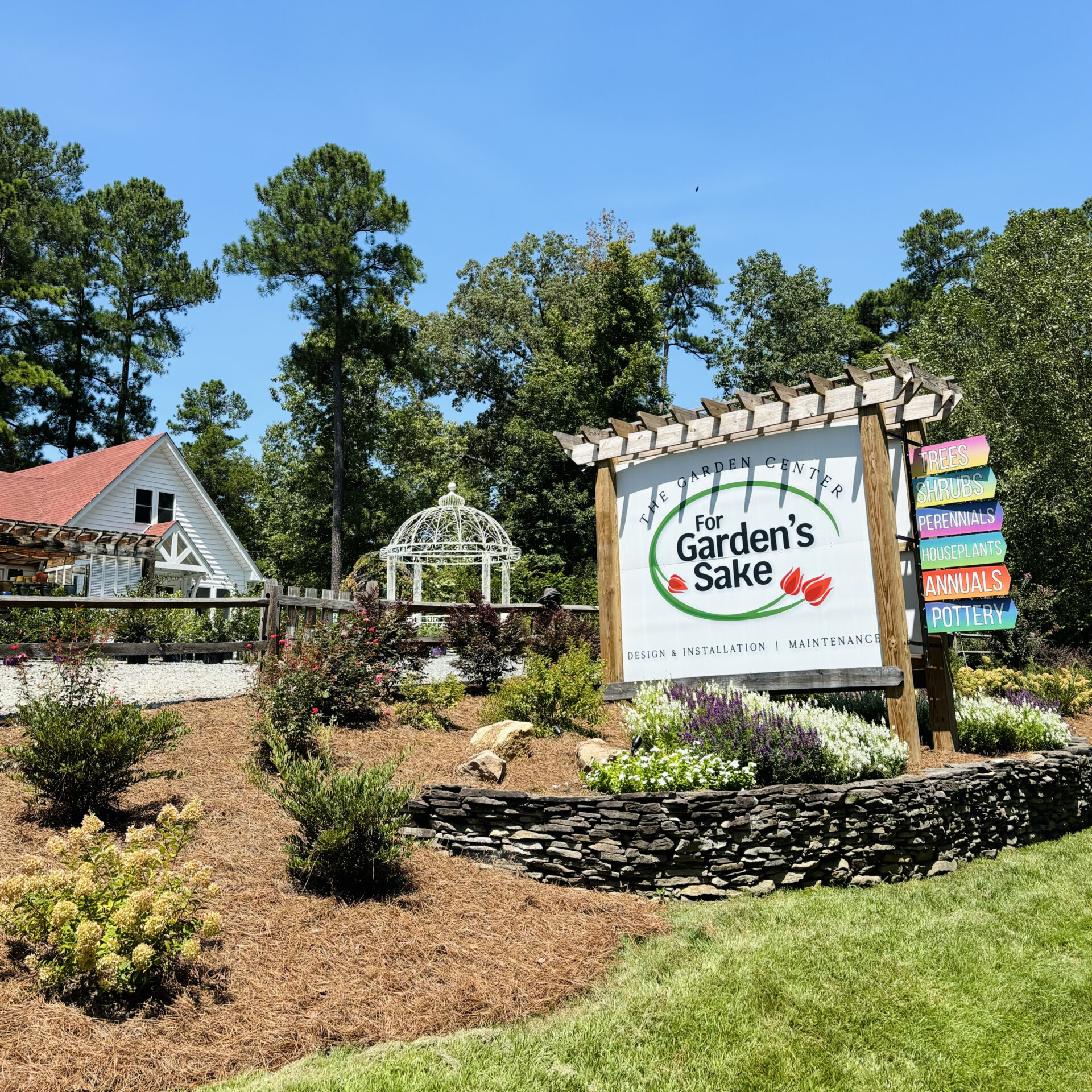 A garden center with a wooden sign displays colorful directions. Nearby, a white house and gazebo sit amidst lush greenery under a clear blue sky.