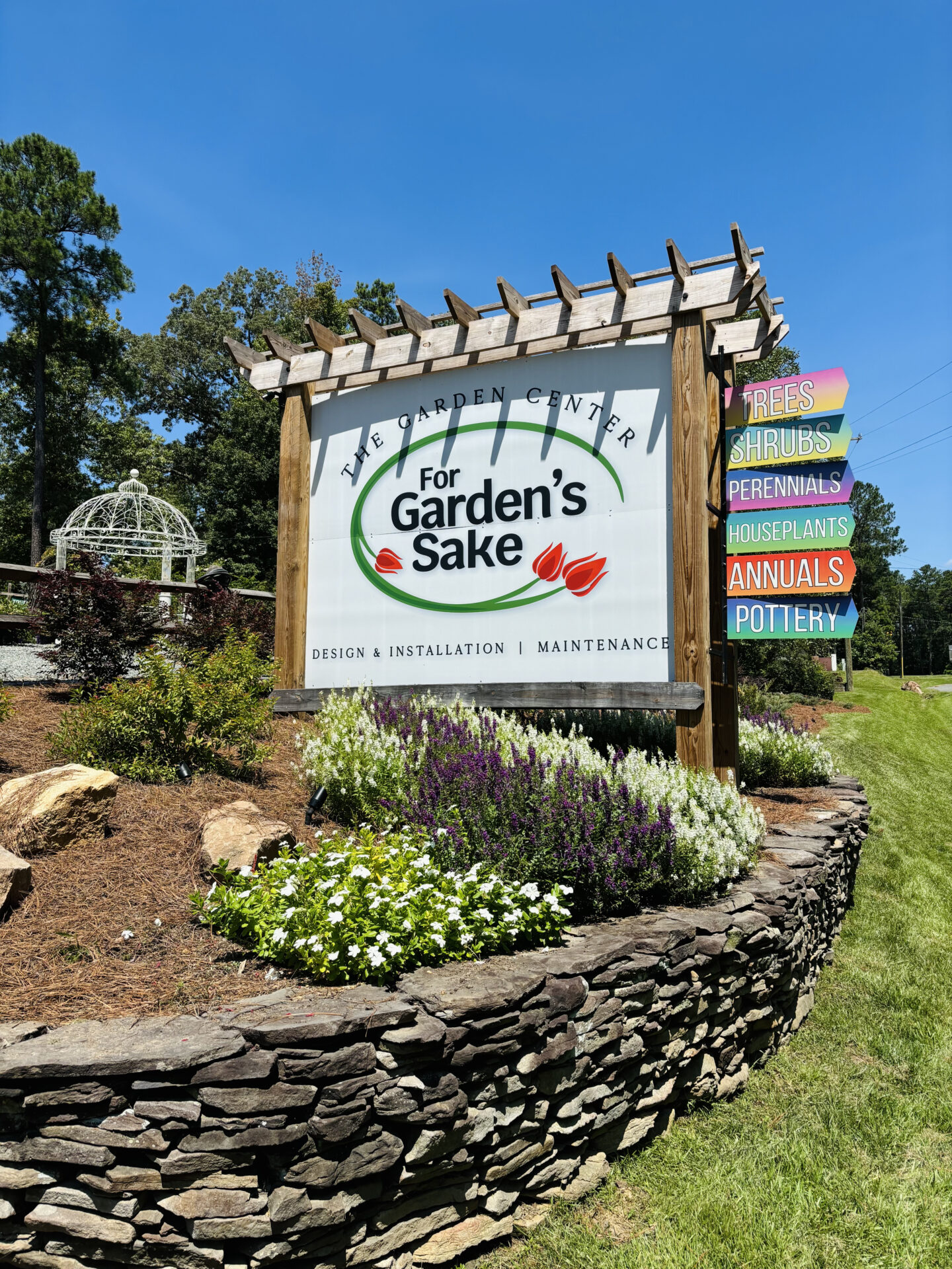 Outdoor garden center sign with colorful flowers, indicating services like design, installation, and maintenance. Surrounded by stone landscaping and greenery.