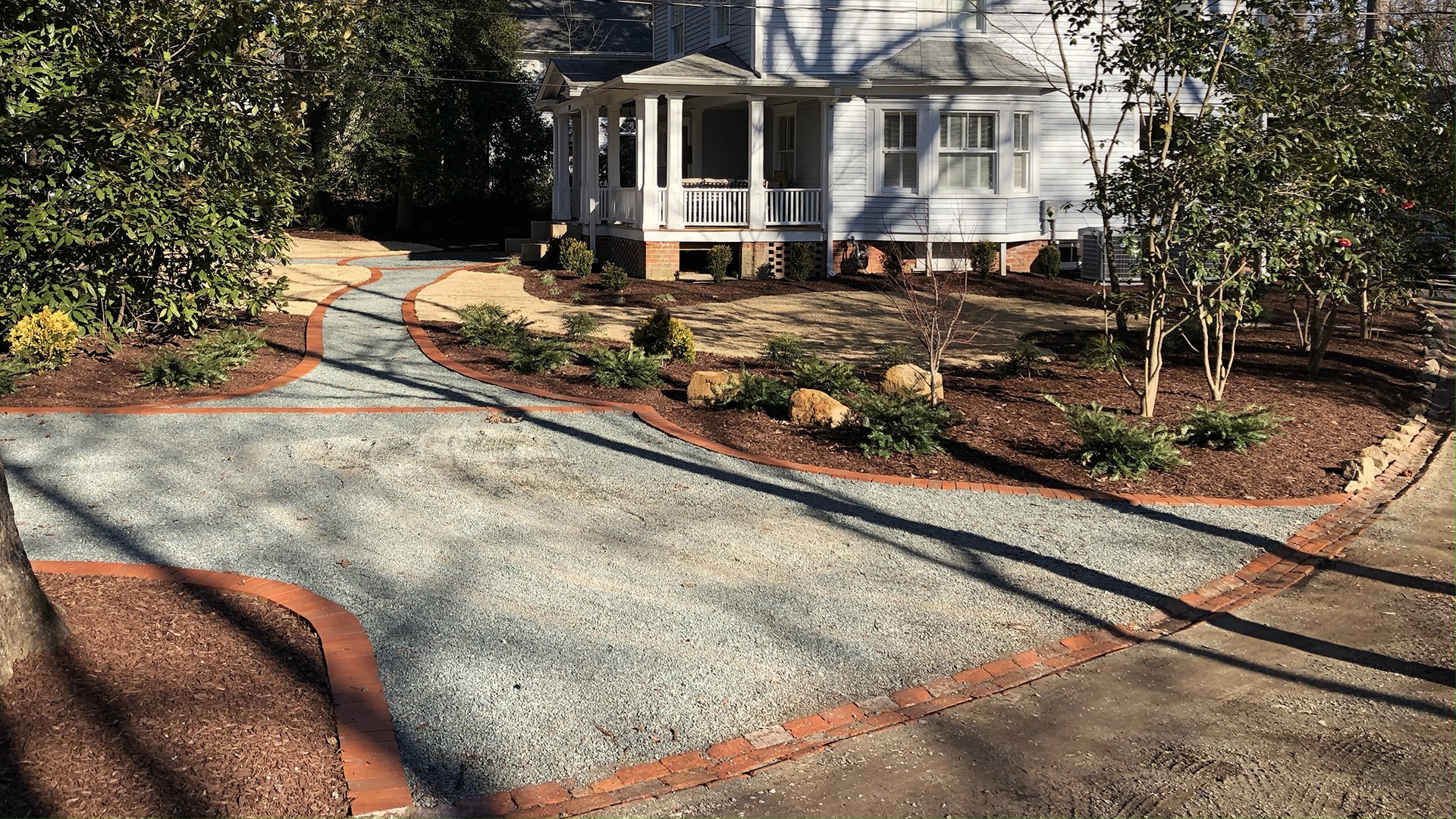 A white house with a porch, surrounded by a landscaped yard featuring a gravel and brick walkway under shadows of nearby trees.