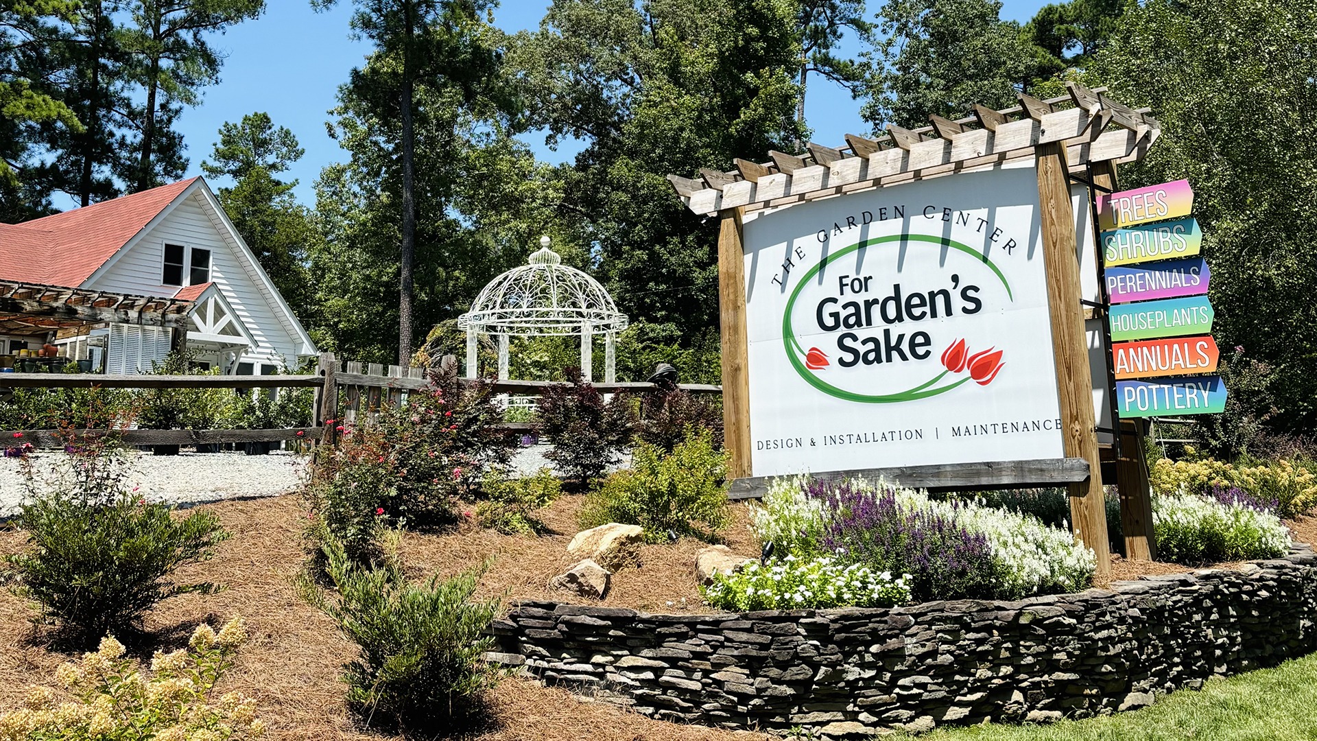 Garden center with a white house, decorative gazebo, and colorful sign listing services like pottery and plants. Surrounded by greenery and trees.