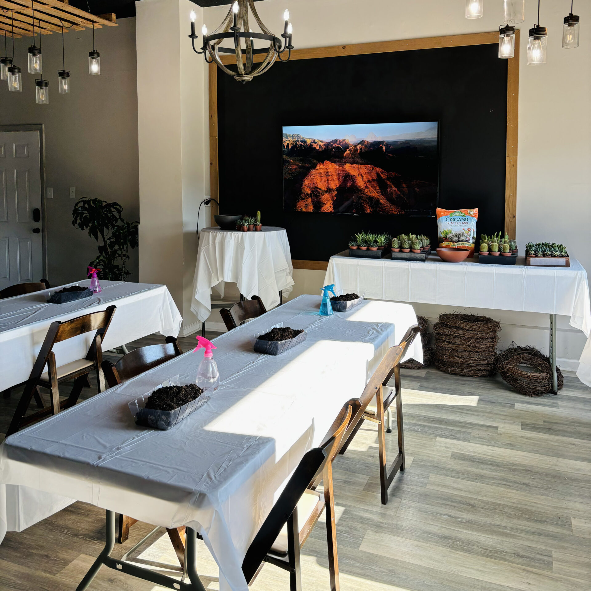 Room with decorated tables, potted plants, and a large photo of a canyon landscape. Chandelier lighting and wooden chairs add a cozy ambiance.