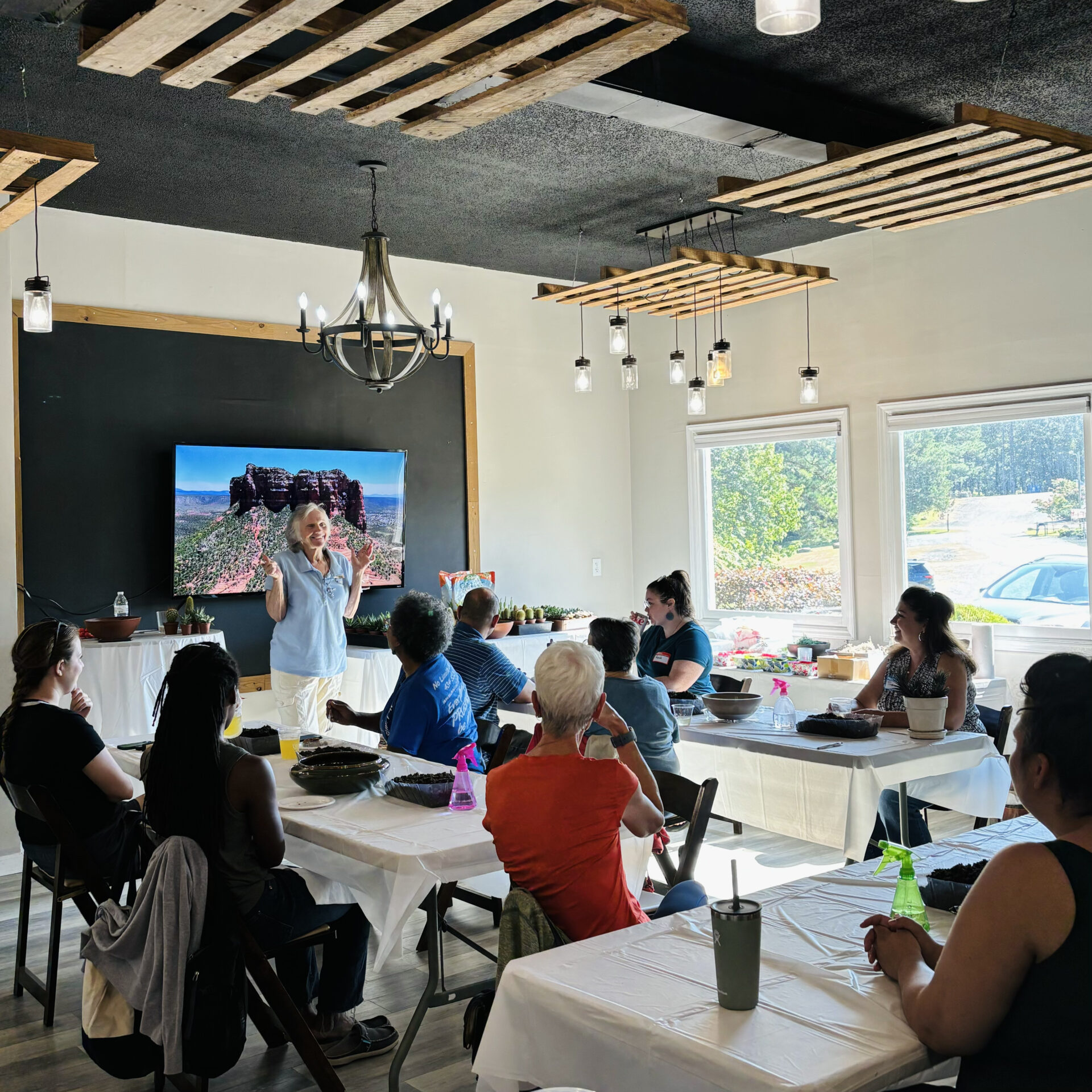 A person speaks to a group in a room with tables and chairs. A large screen displays an outdoor image.