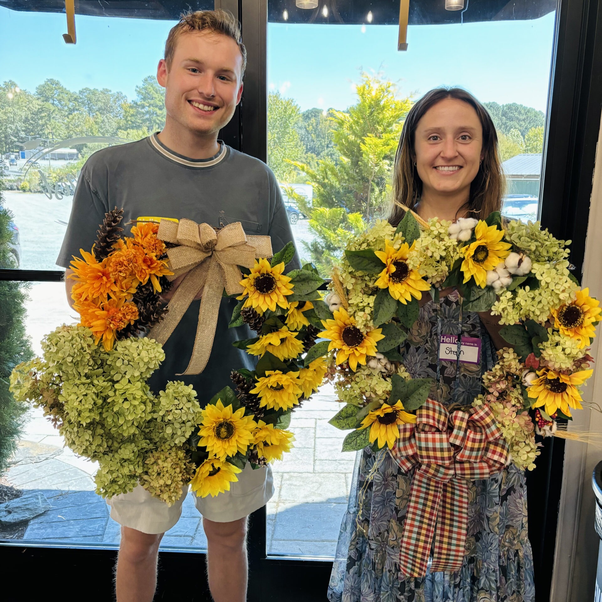 Two people holding decorative sunflower wreaths stand in front of large windows, with greenery and trees visible outside.