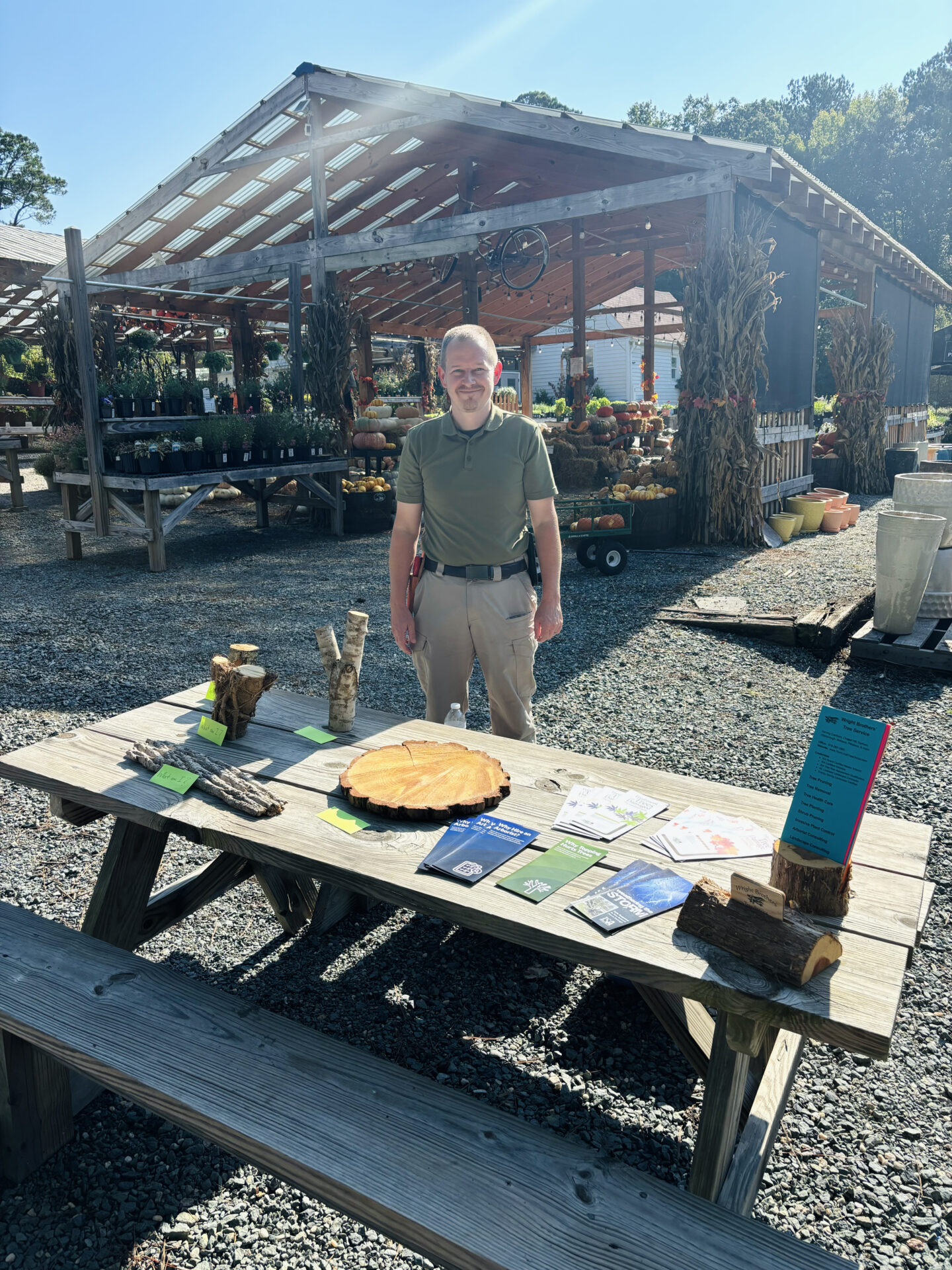 A person stands by a display table outdoors at a plant nursery. The table features brochures and a wooden decor piece under a shaded structure.