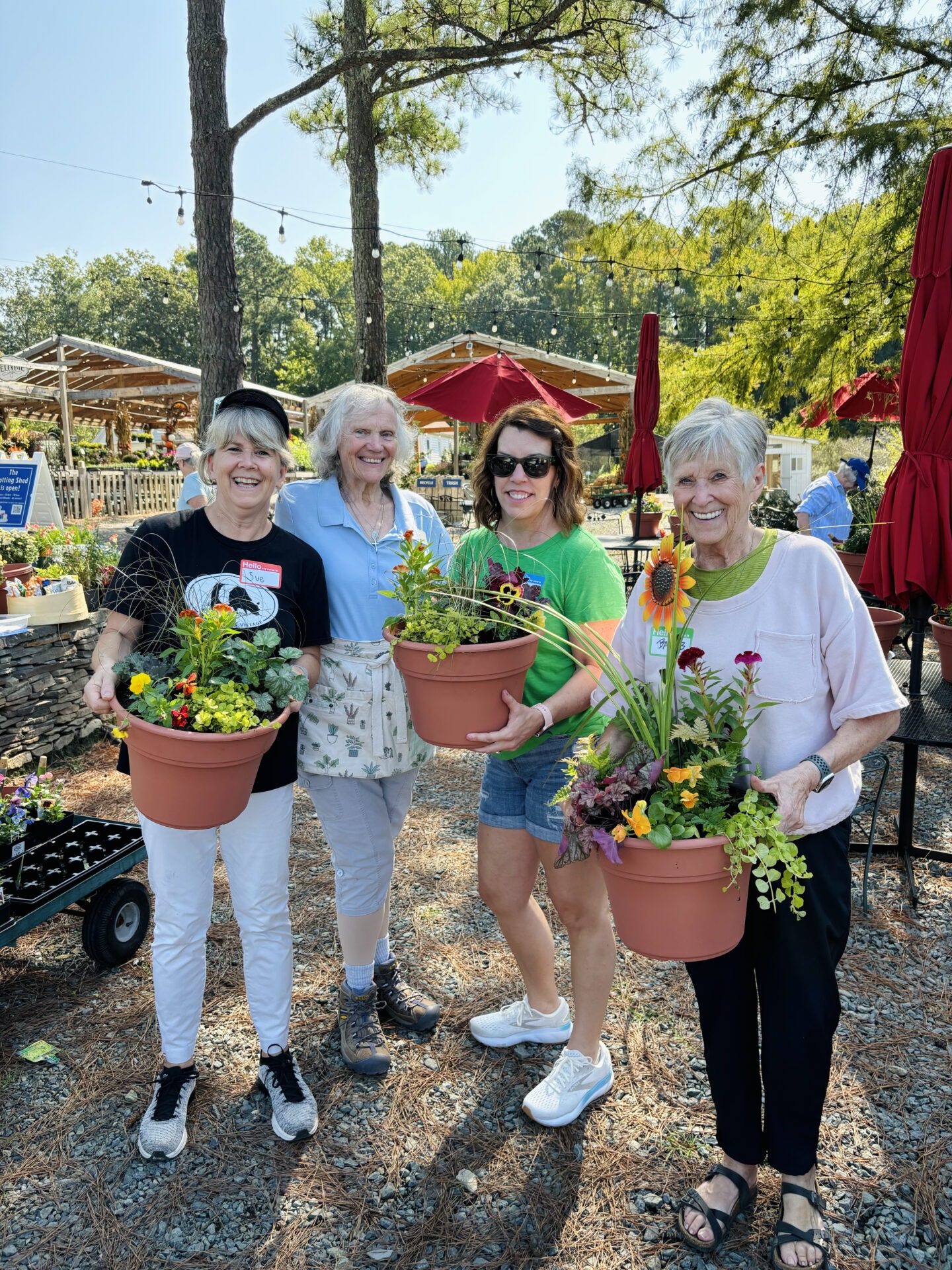 Four people smiling, holding flower pots at an outdoor garden center. Red umbrellas and green trees in the background enhance the vibrant scene.