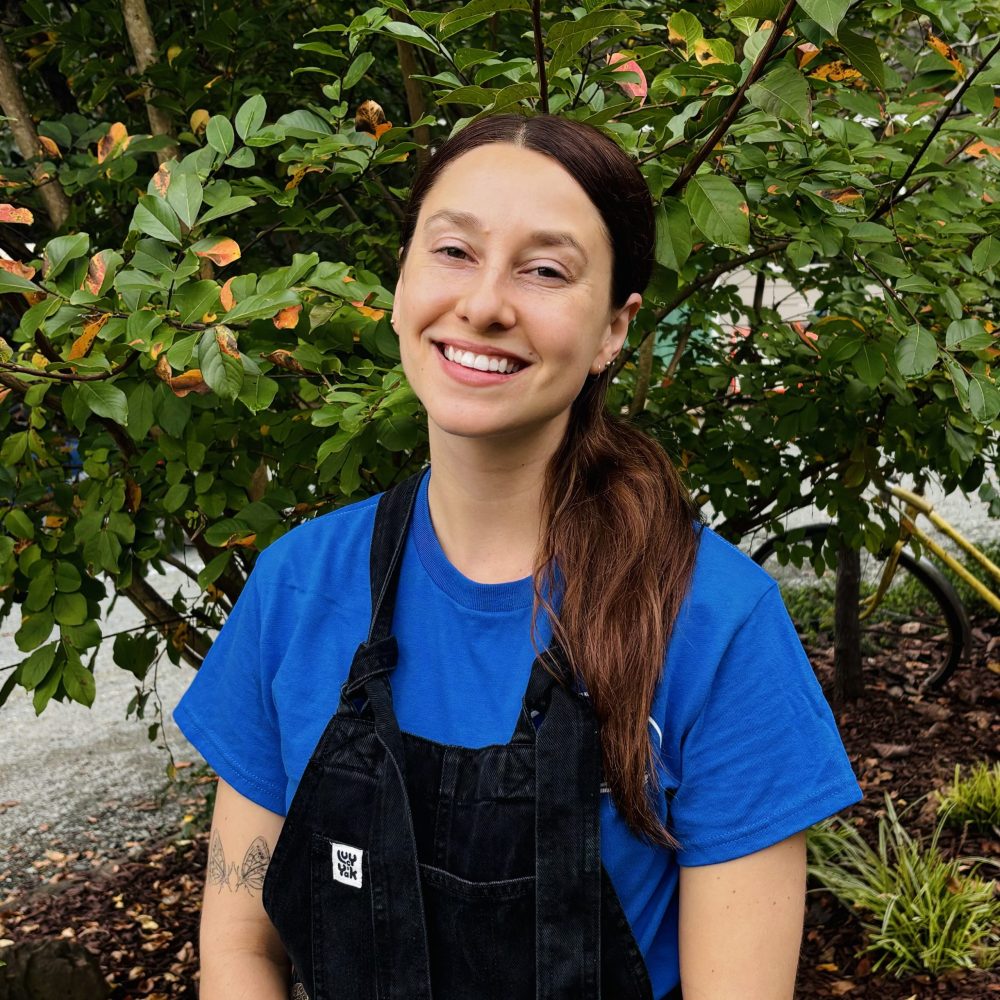 A person in a blue shirt and black overalls is smiling outdoors with greenery in the background. No recognizable landmarks or buildings are visible.