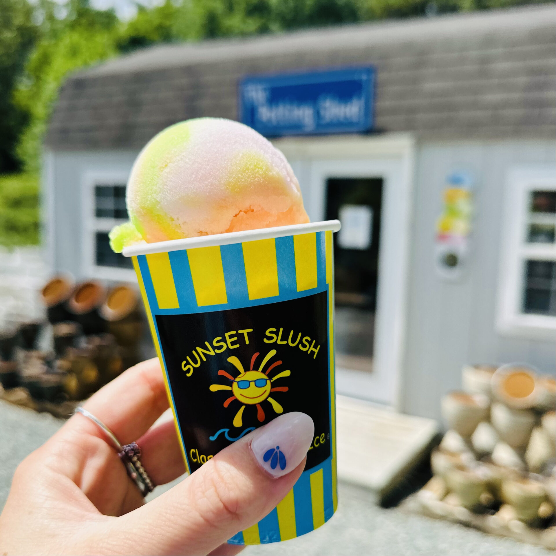 A person holds a colorful slushie in front of a small building with a sign, surrounded by logs and greenery.