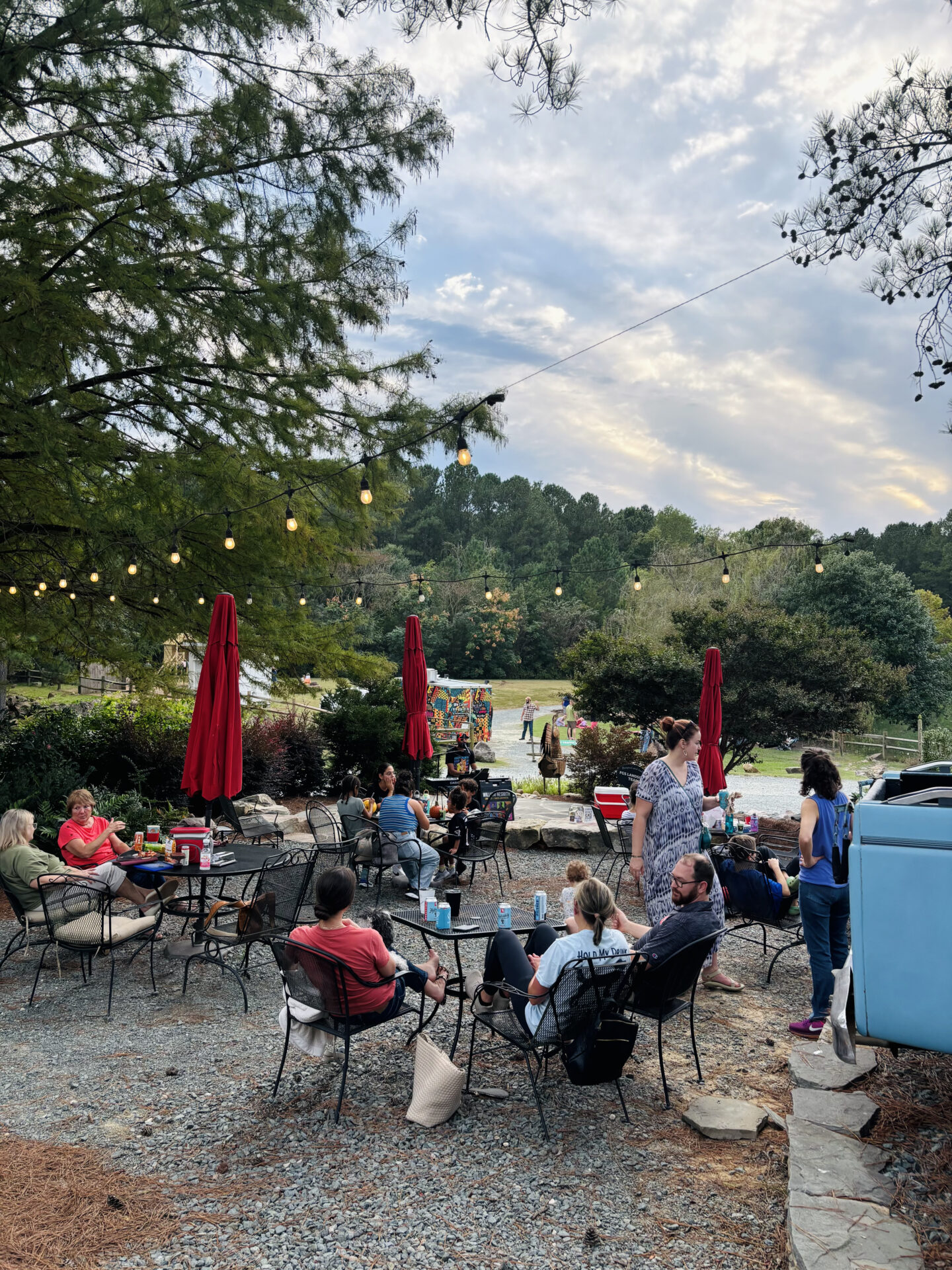 A group of people relax outdoors on a patio with string lights and red umbrellas, surrounded by lush greenery under a cloudy sky.