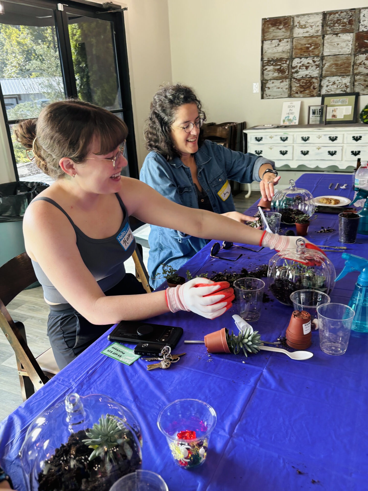 Two people smile while working on terrarium projects at a table with plants, soil, and tools. Bright, well-lit room with natural decor.