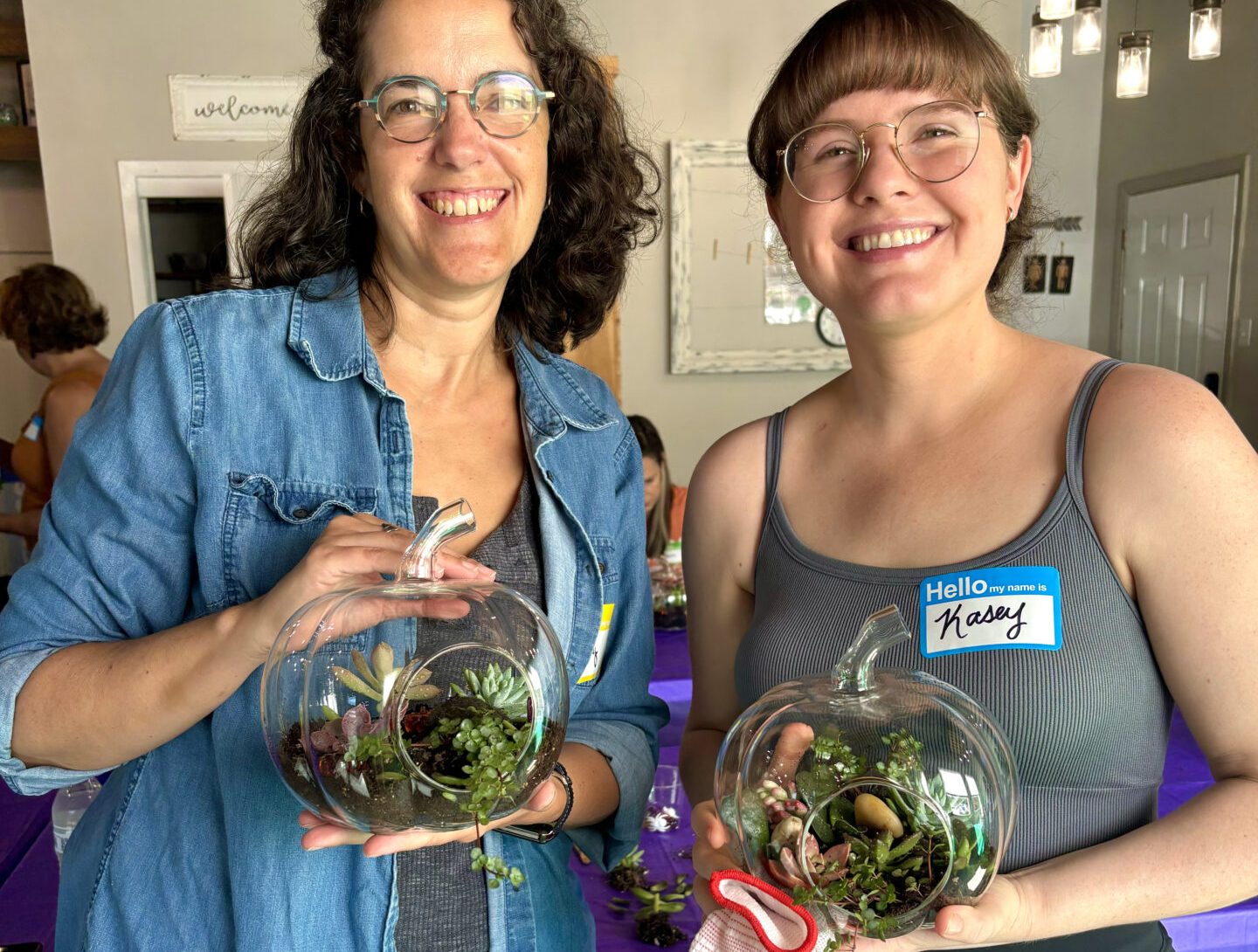 Two people indoors, smiling and holding glass terrariums with plants. They stand beside a table with a purple tablecloth in a cozy setting.