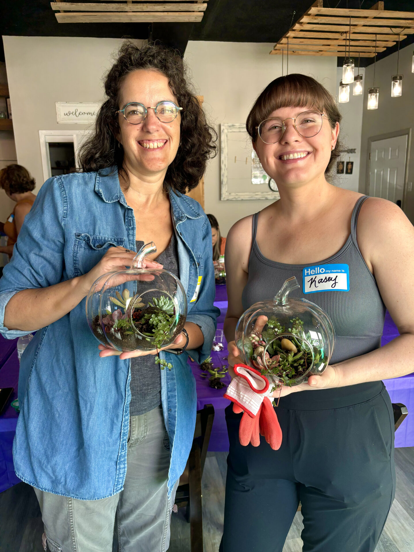 Two people indoors, smiling and holding glass terrariums with plants. They stand beside a table with a purple tablecloth in a cozy setting.