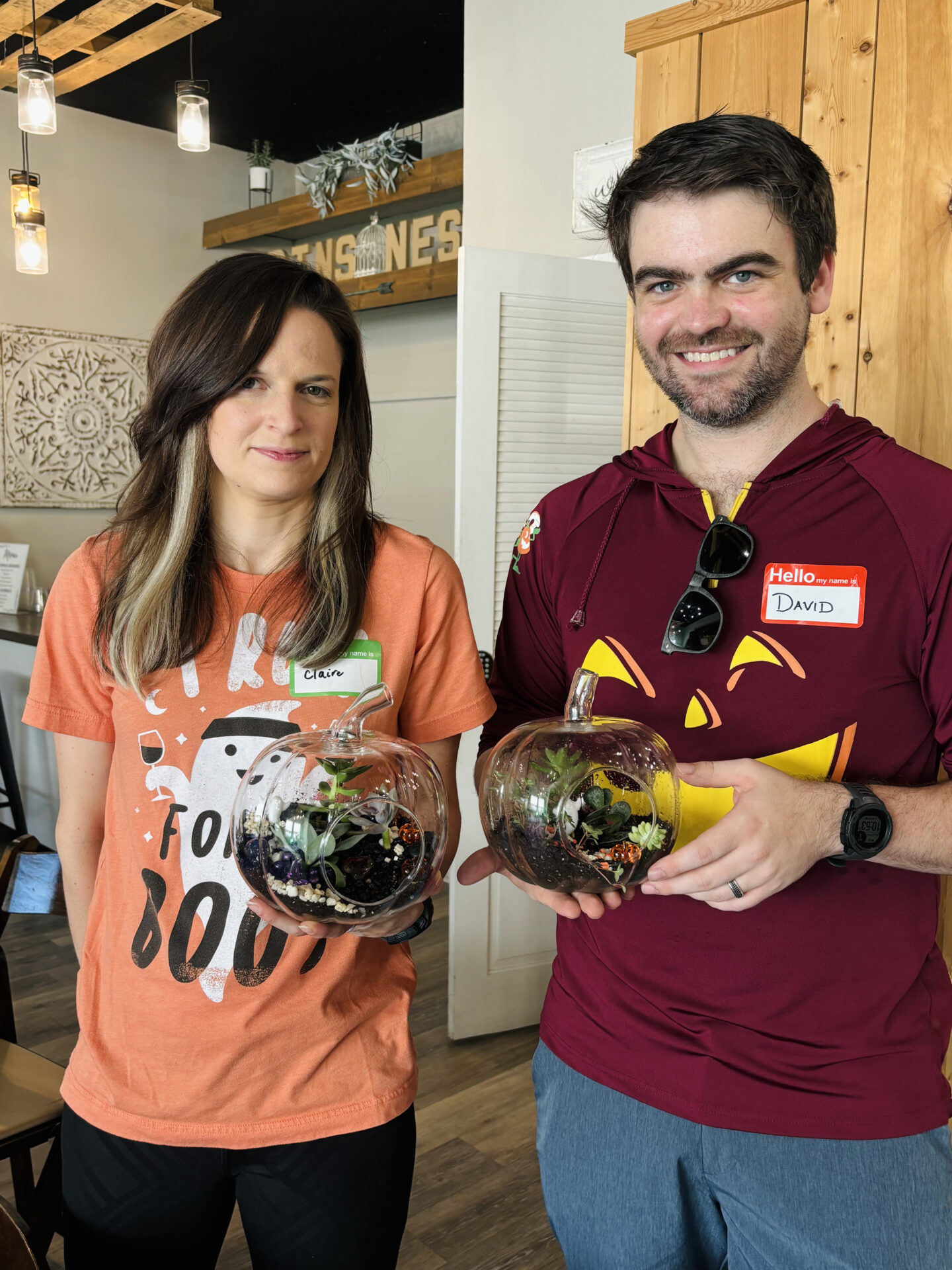 Two people in casual attire hold glass pumpkin terrariums inside a cozy room with wooden decor and soft lighting.