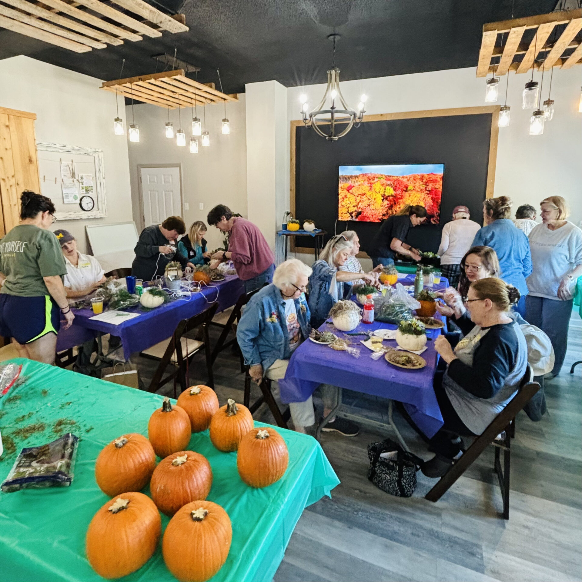 A group of people engage in a pumpkin-decorating activity indoors, surrounded by tables, chairs, and hanging lights creating a cozy atmosphere.