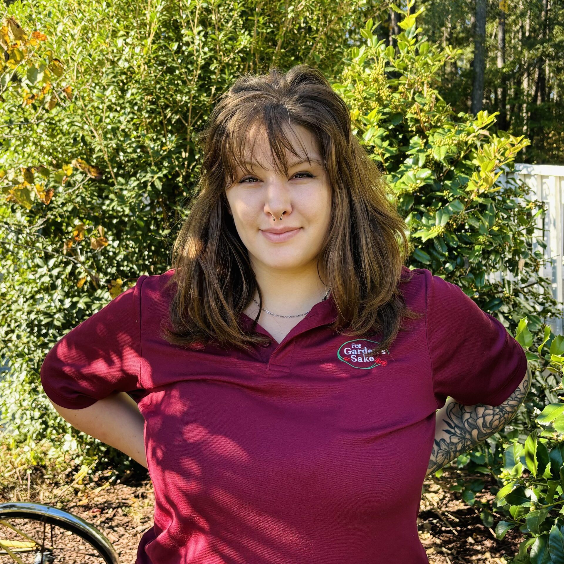 A person in a red shirt stands outdoors, surrounded by green bushes and trees, smiling under the sunlight. Tattoo visible on arm.