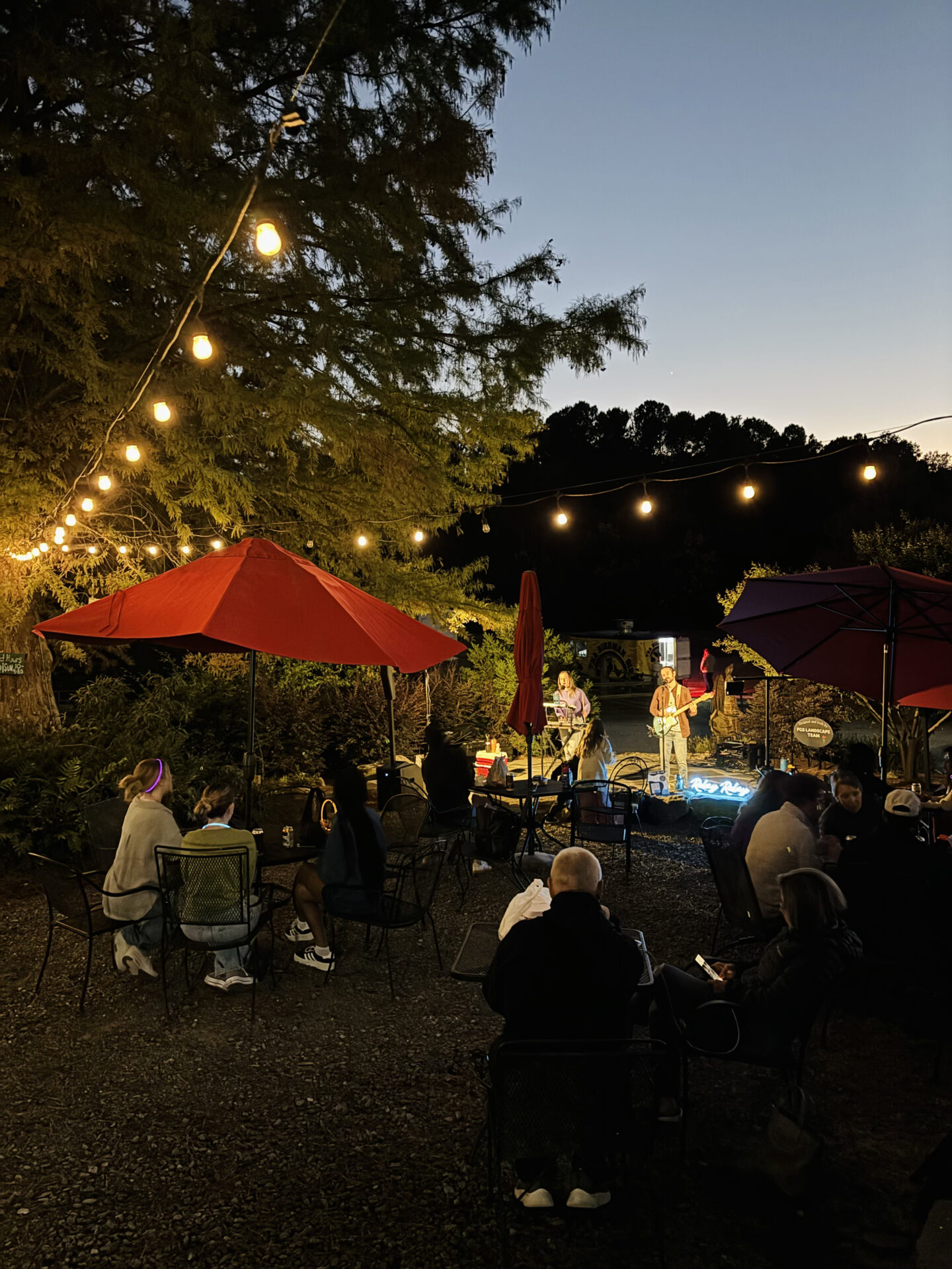 Evening outdoor music performance with string lights and red umbrellas. Audience seated at tables, listening under a twilight sky, surrounded by trees.