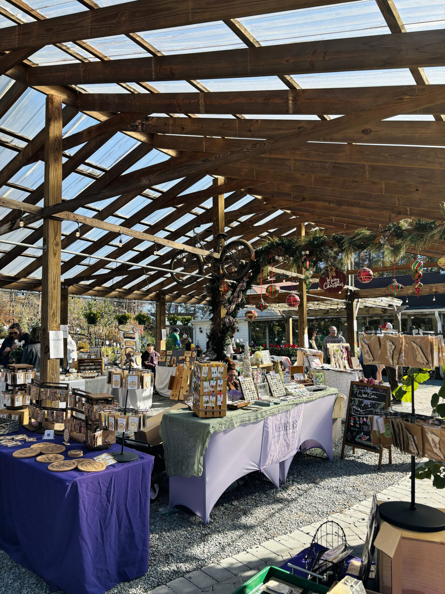 Outdoor market scene with wooden stalls displaying crafts and artwork. People browse under a wooden roof adorned with festive decorations. Bright, sunny day.