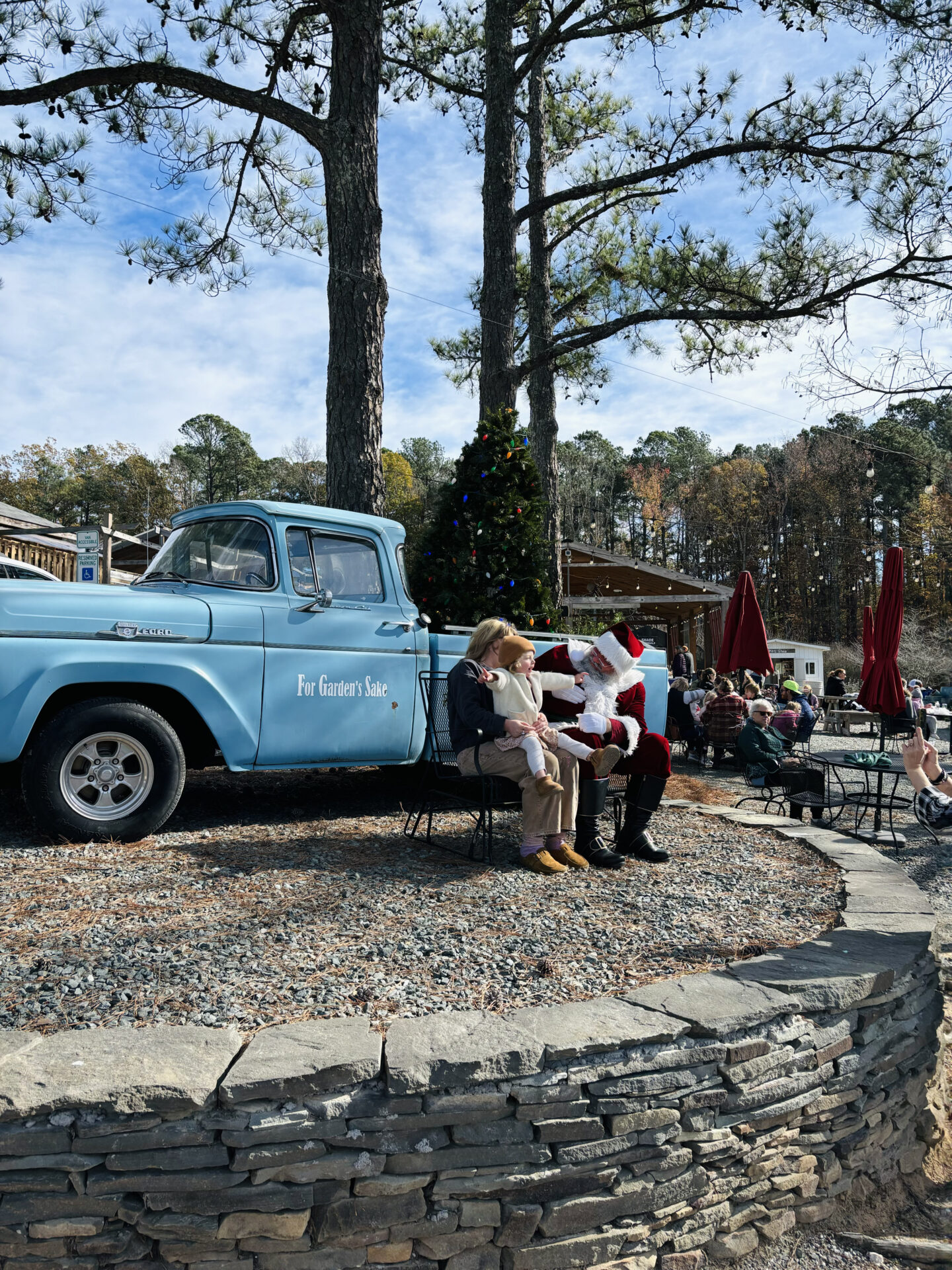 A person and child sit with Santa near a vintage blue truck outside surrounded by trees and people, with red umbrellas in the background.