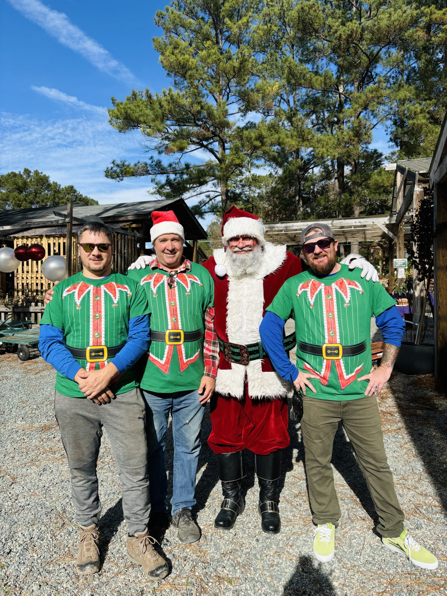 Four people, including one dressed as Santa, pose outside wearing festive shirts. The background features trees and a wooden structure.