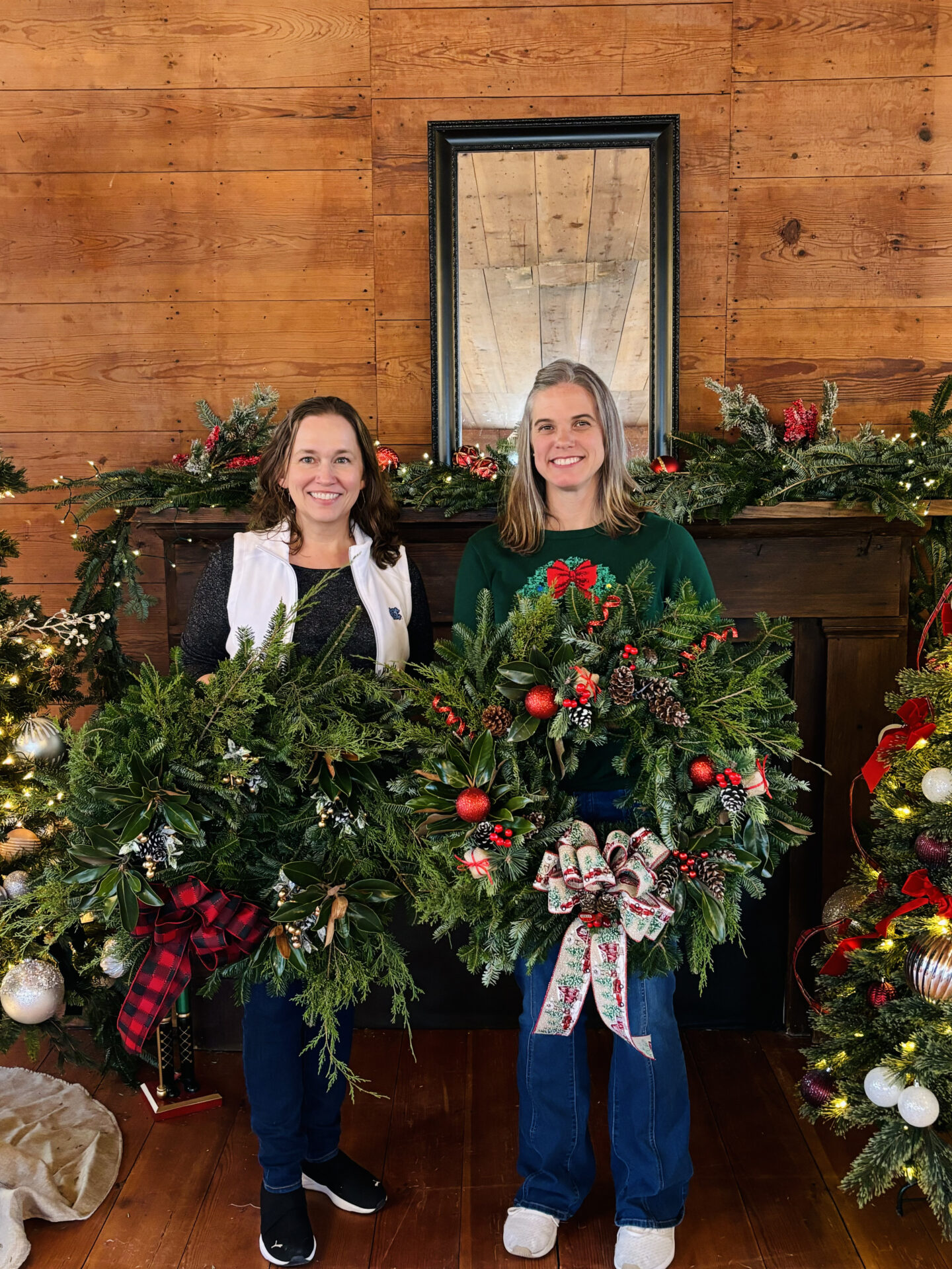 Two people holding festive wreaths stand indoors with decorated fireplace and mirror in background. Holiday decorations include greenery, ribbons, and ornaments.