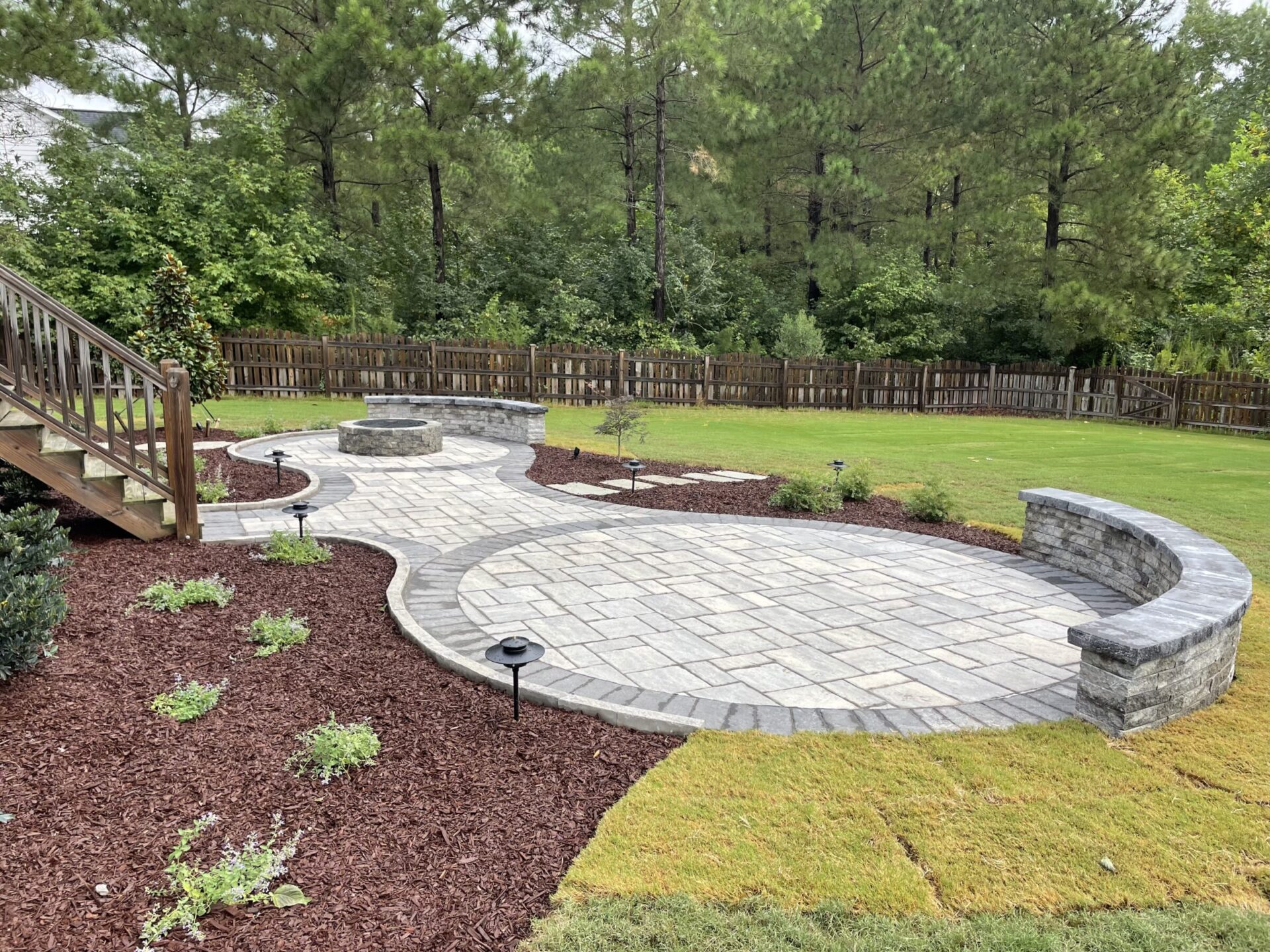 A backyard features a stone patio with circular seating area, wooden stairs, fresh landscaping, grass, mulch, and surrounding trees and fencing.