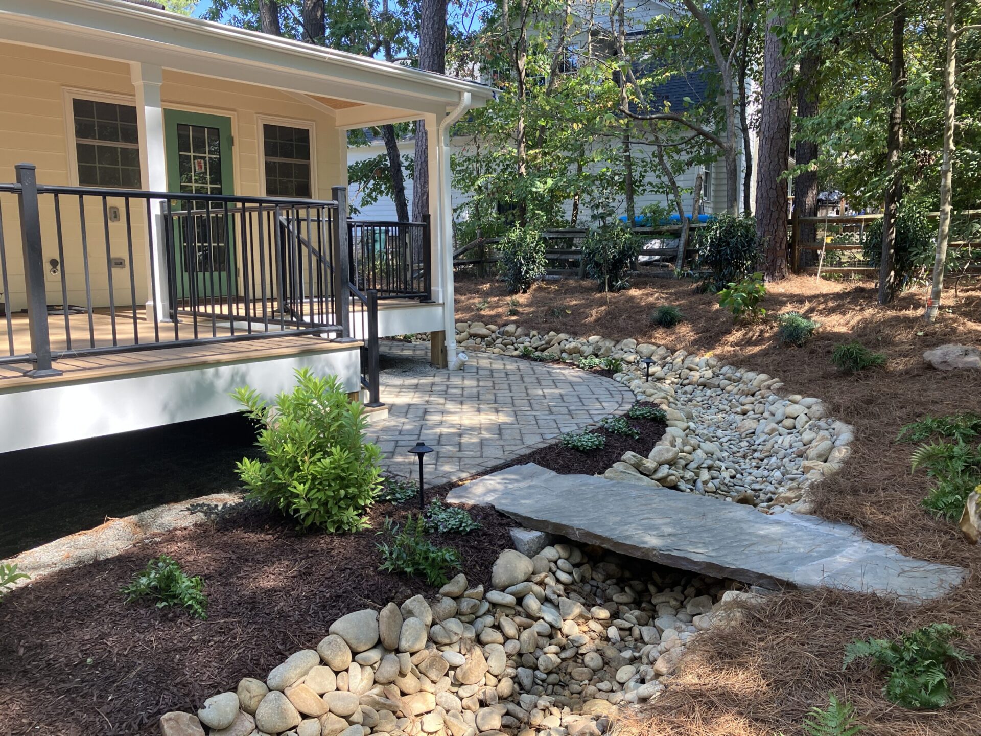 A backyard with a wooden deck, stone path, dry creek bed, and lush greenery, surrounded by trees and fenced by a wooden barrier.