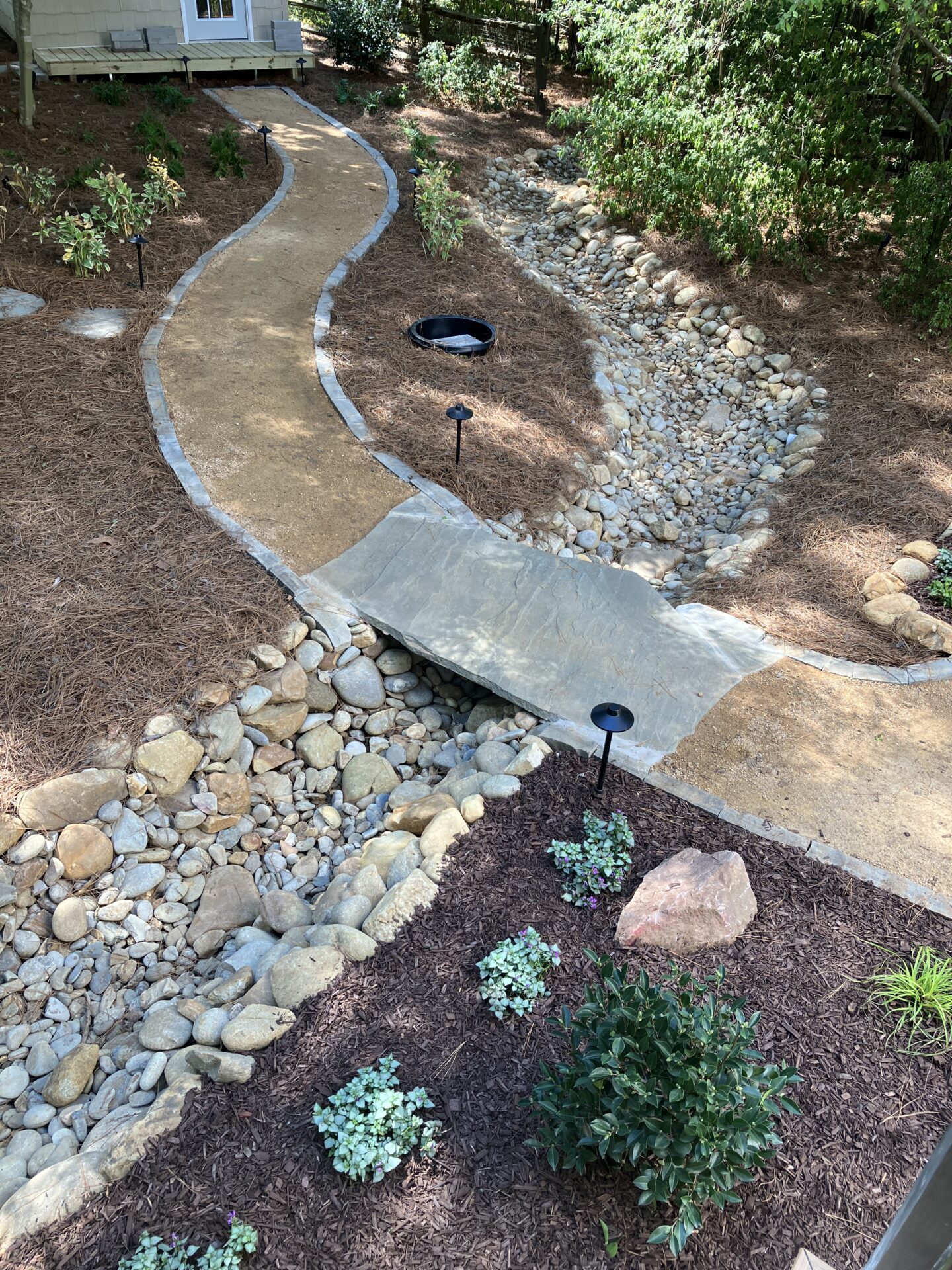 A serene garden pathway features a stone bridge over a dry creek bed, surrounded by neatly arranged plants and illuminated by small lamps.