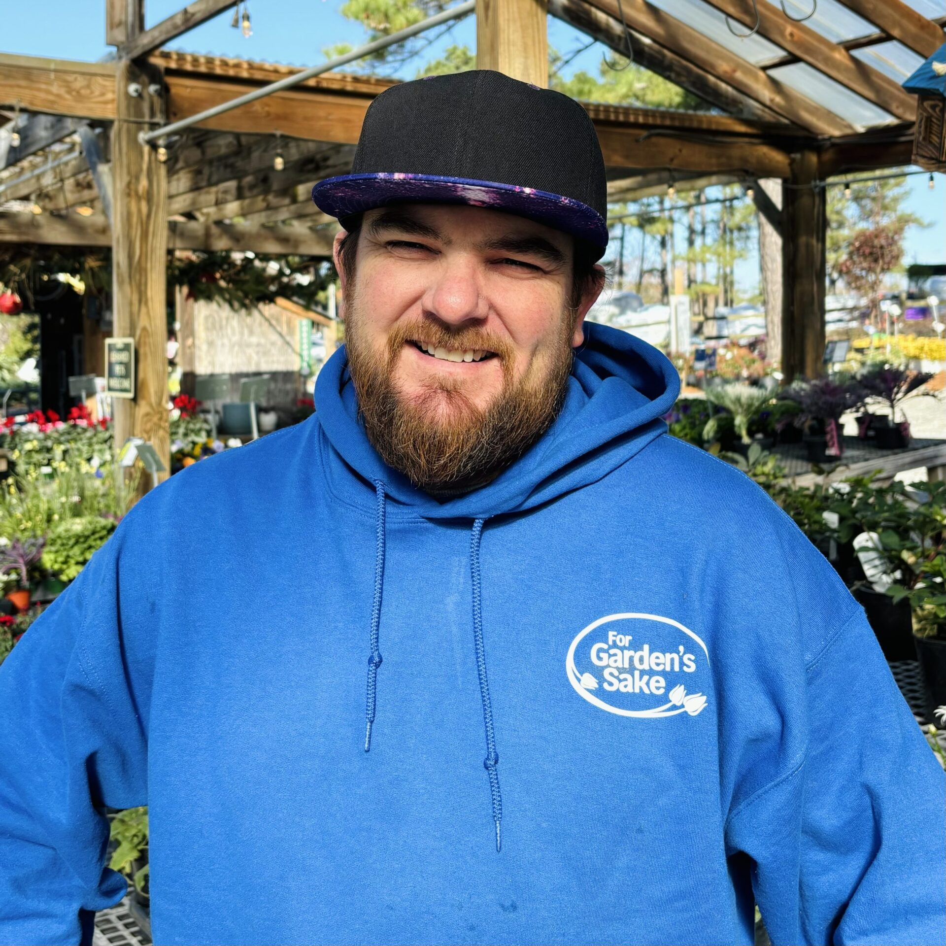 A person in a blue hoodie and cap stands smiling among plants in a garden center with a wooden structure above.