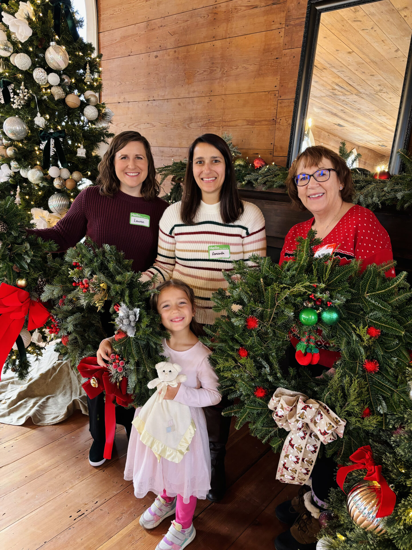 Four people indoors with festive wreaths and a decorated Christmas tree in the background. Smiling child holds a plush toy.