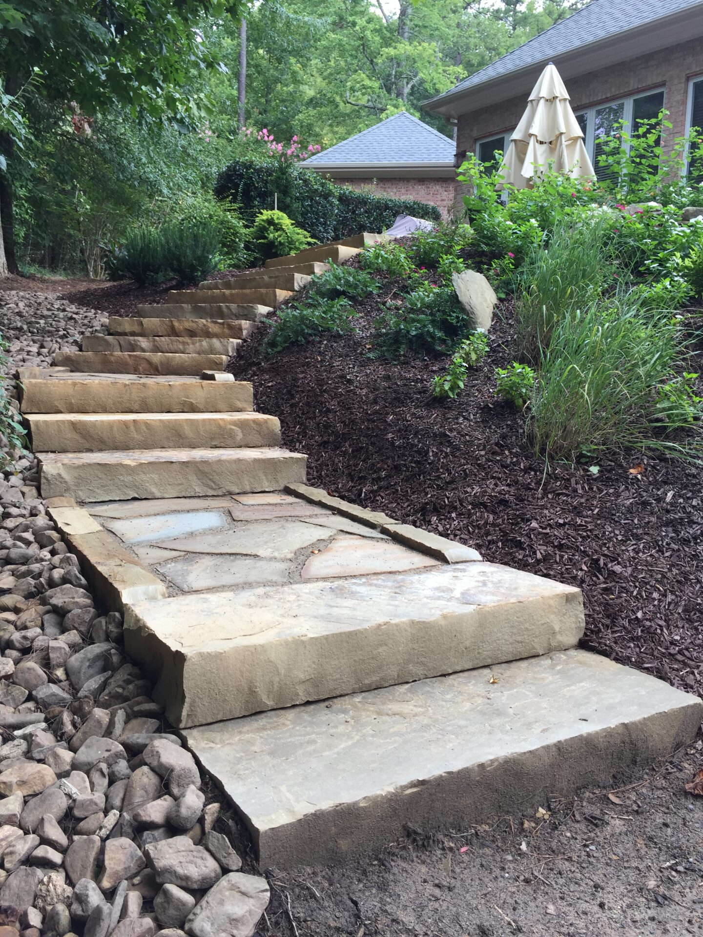 Stone steps lead up through a landscaped garden, surrounded by lush greenery and mulch. A building with an umbrella is visible.