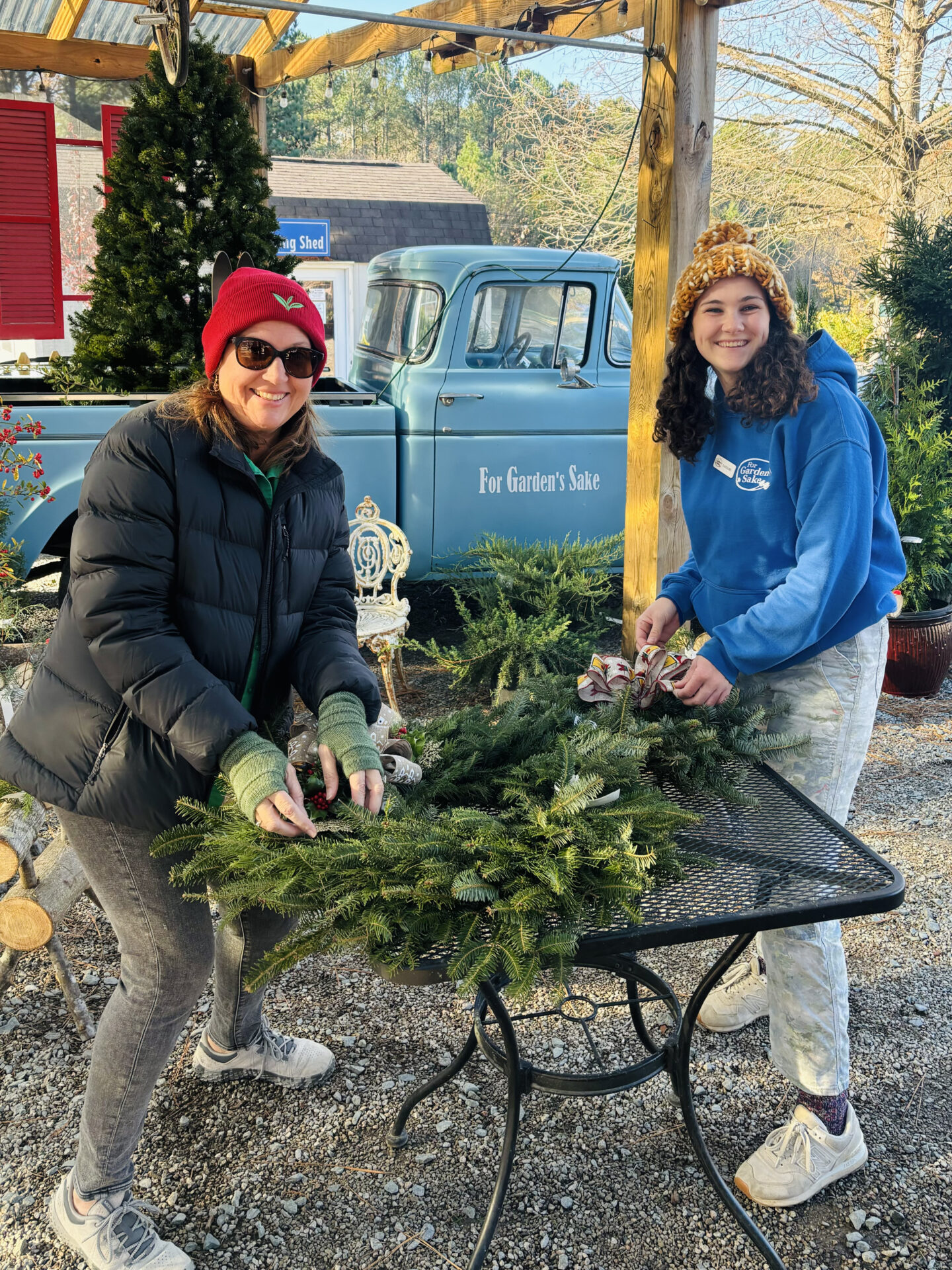 Two people crafting holiday wreaths outdoors near a vintage blue truck, surrounded by greenery and festive decorations under a wooden pergola.