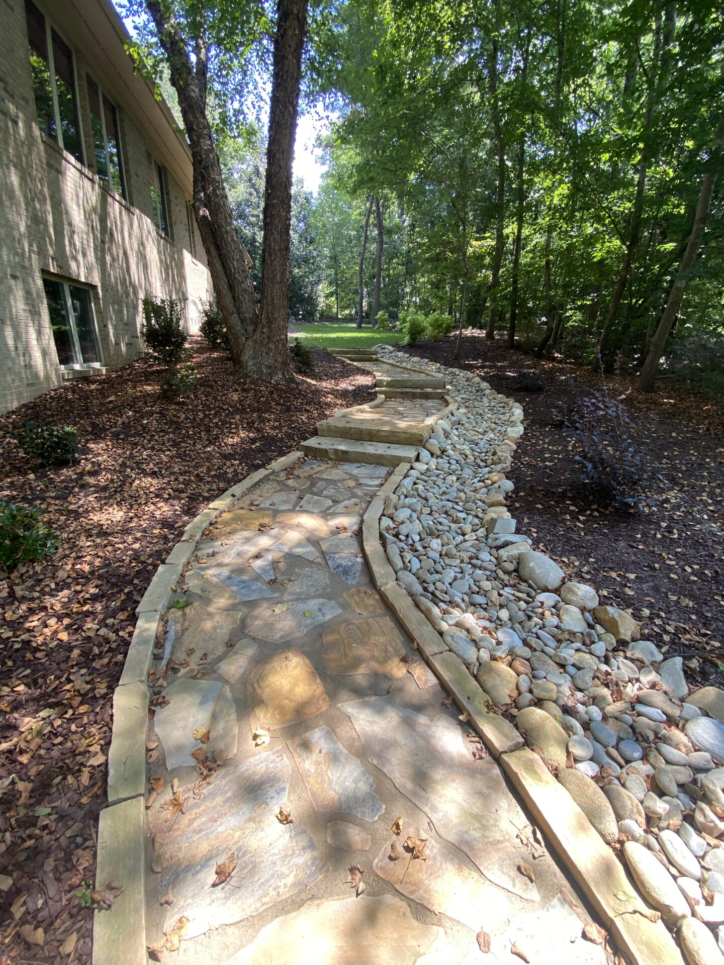 Stone path winds through a wooded garden beside a building. Leafy trees provide shade, and small stone border accents the pathway's edge.