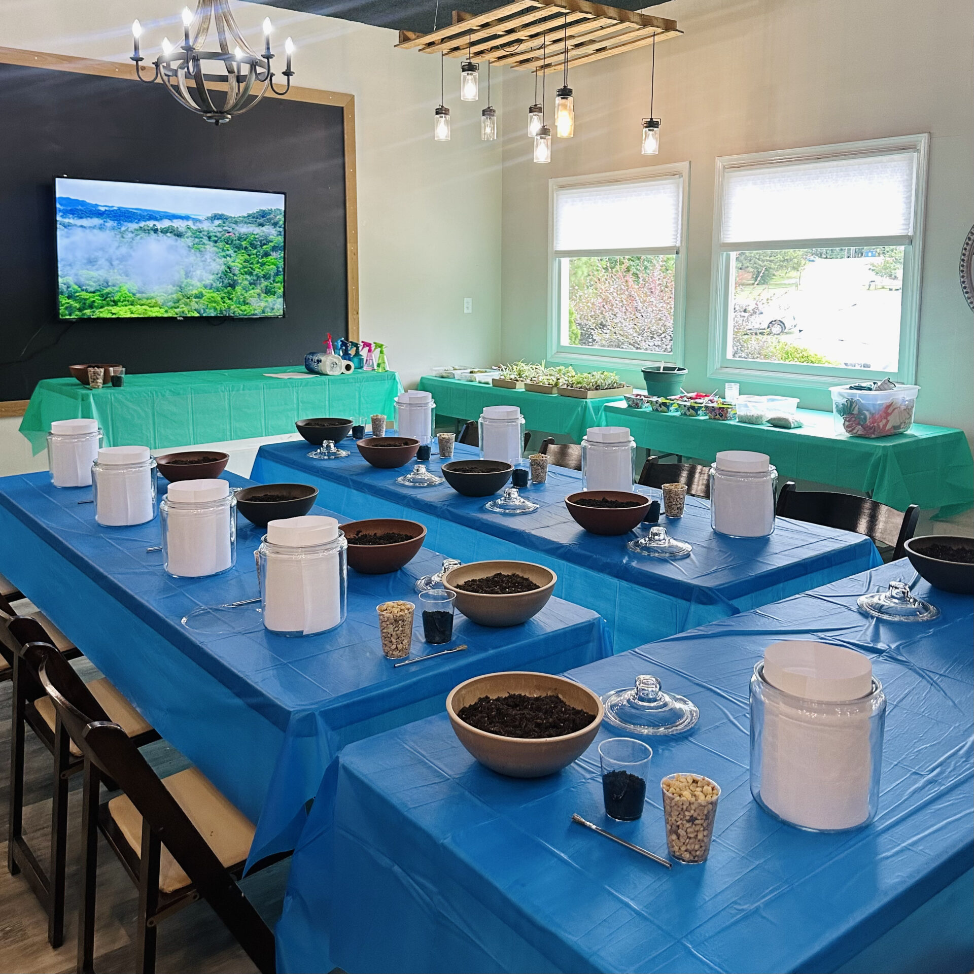 Room set for a workshop with blue tablecloths, jars, bowls, and ingredients. A landscape image on the screen, large windows, and lighting fixtures.