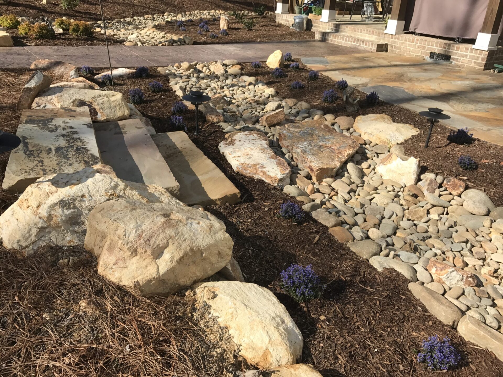 A landscaped garden with large rocks, pebbles, and small purple flowers. A paved pathway and stone building are in the background.