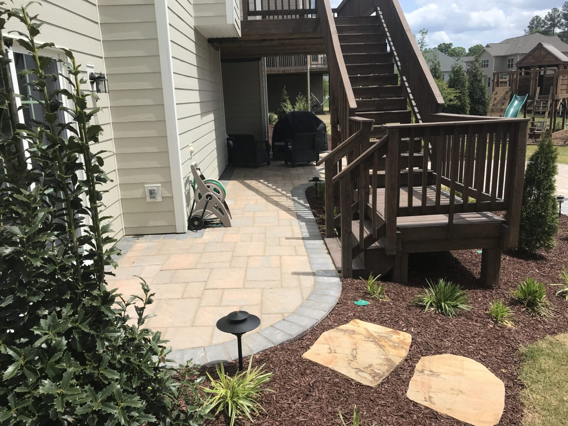 A backyard patio with wooden stairs, stone path, and garden area. Visible houses and a playground in the background under a cloudy sky.