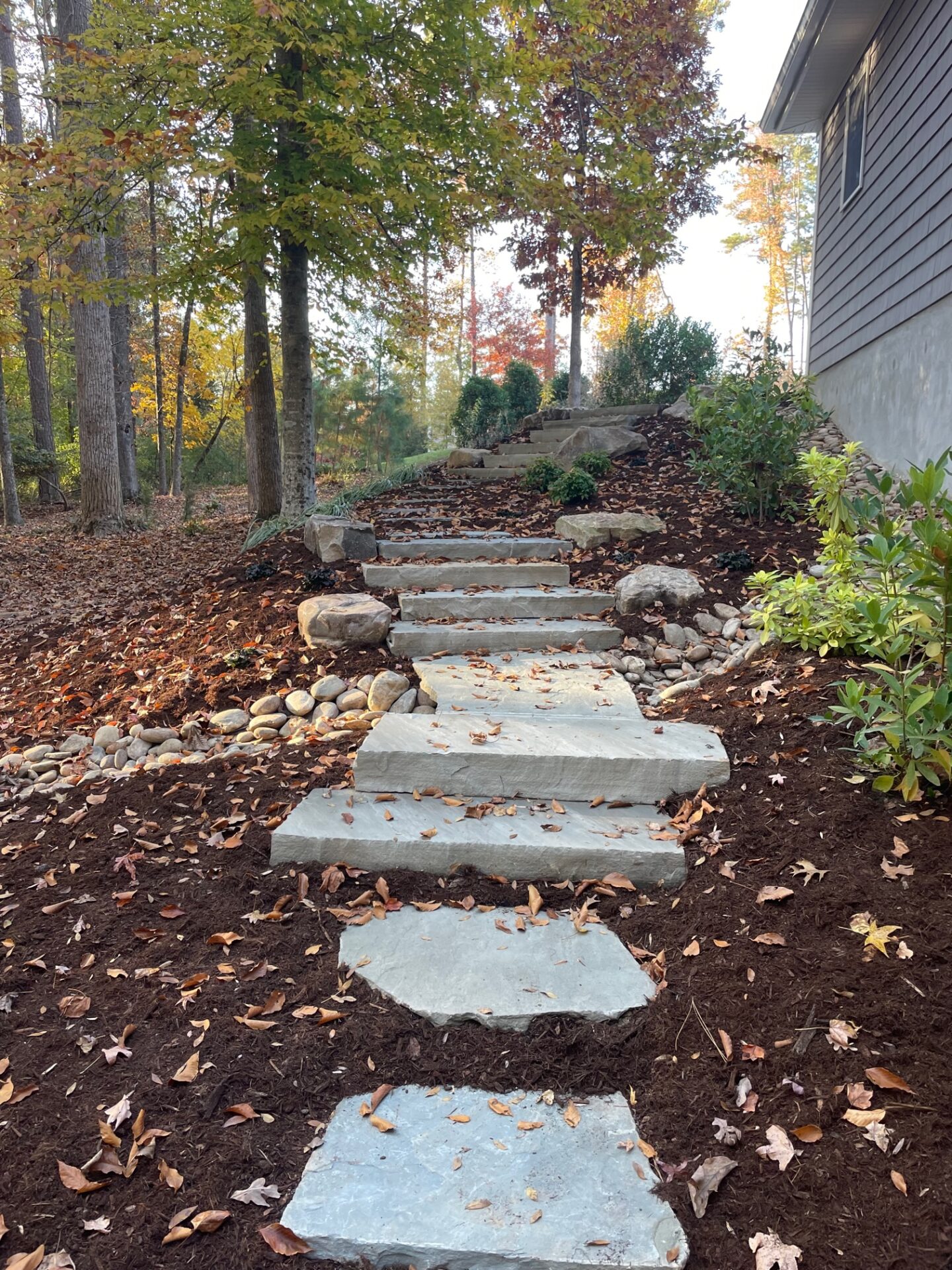 A stone path ascends through a wooded garden featuring trees with autumn leaves and a house on the right.