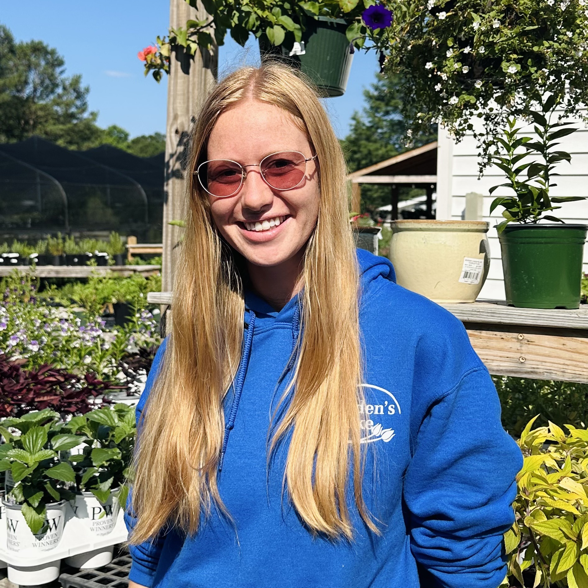 A person in sunglasses and a blue hoodie stands in a garden center, surrounded by plants and pots, under a sunny sky.