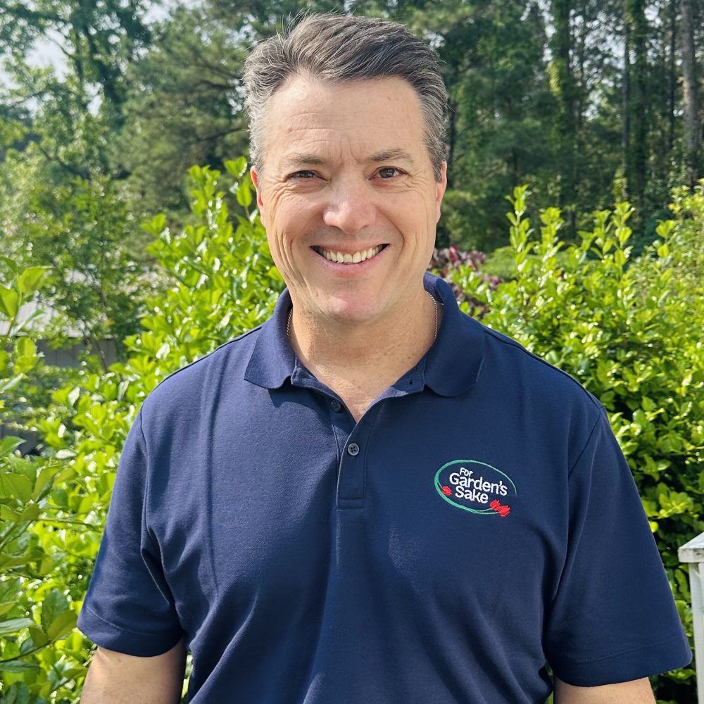 A person in a navy polo shirt stands smiling in front of lush greenery. The backdrop consists of dense trees in a sunny outdoors setting.