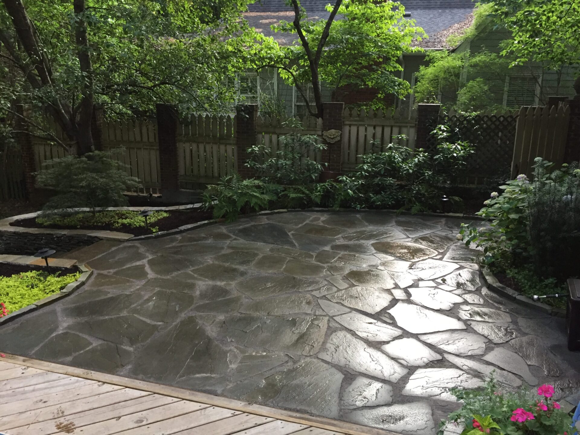 A tranquil backyard with a flagstone patio, lush greenery, and wooden fencing. Trees provide shade, creating a peaceful, natural ambiance.