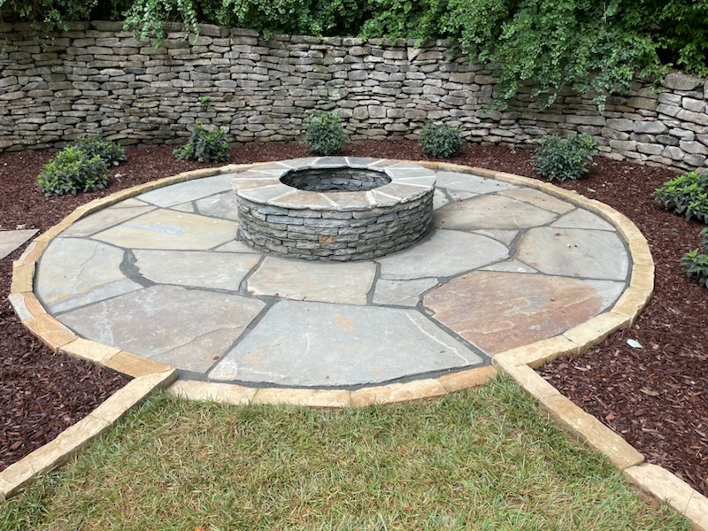 A circular stone fire pit is surrounded by a paved patio, bordered by mulched areas with small shrubs, and backed by a stone wall.