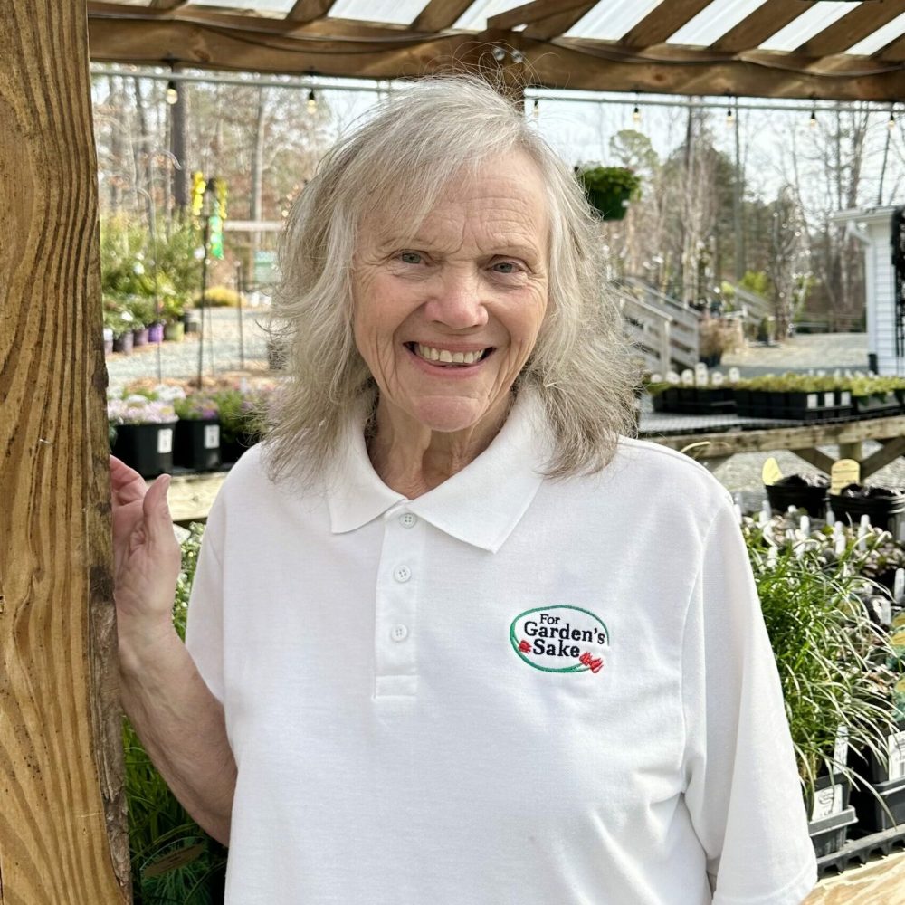 A person in a garden center wearing a white polo shirt with "For Garden's Sake" logo, surrounded by plants and wooden structures.