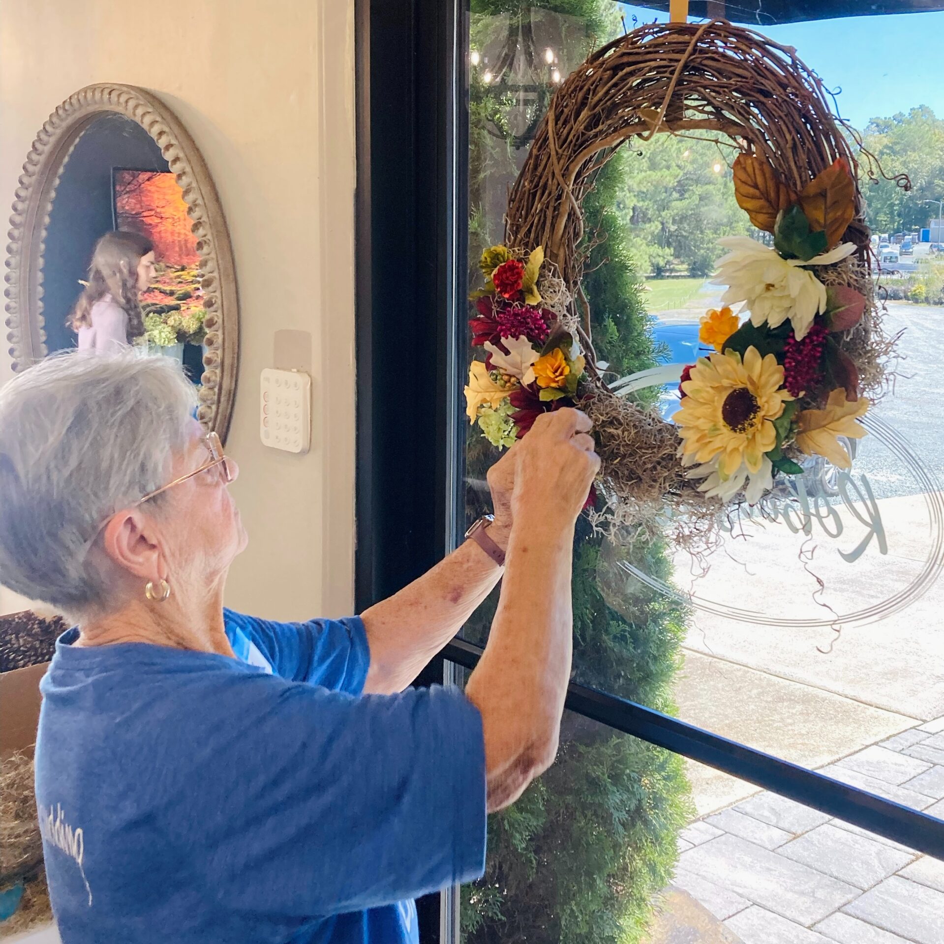 A person hangs a floral wreath on a glass door. Natural light highlights the decorative elements. A mirror and outdoor view are visible.