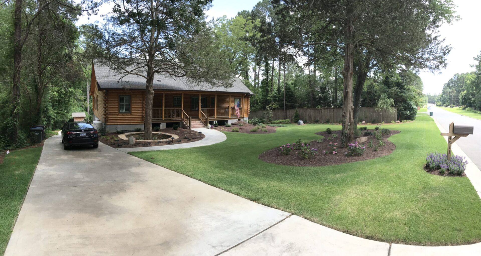 A wooden house with a front porch surrounded by a well-maintained lawn and trees. A car is parked in the driveway.