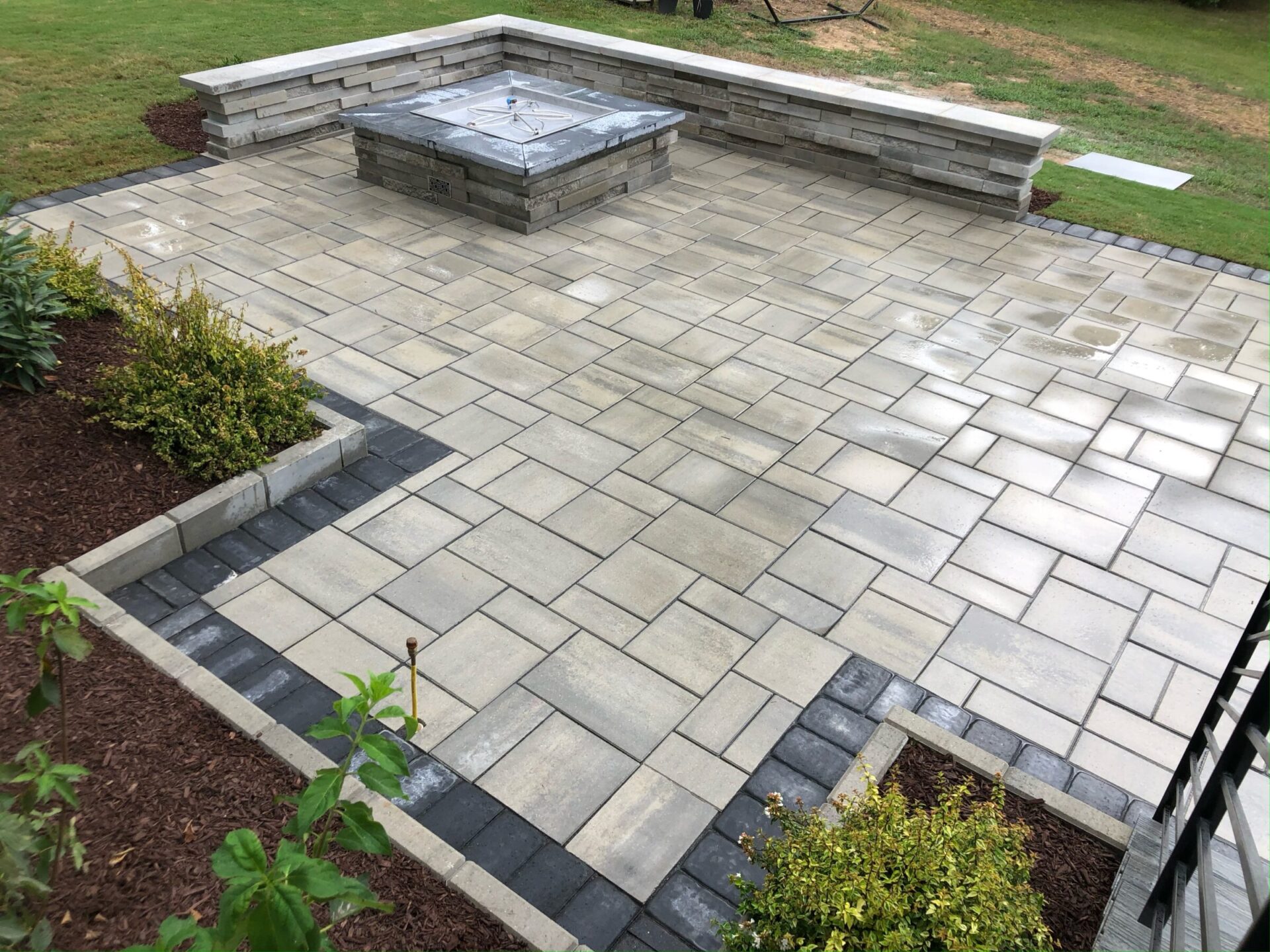 A modern outdoor patio with stone paving and a central fire pit, surrounded by low brick walls and landscaped with shrubs.