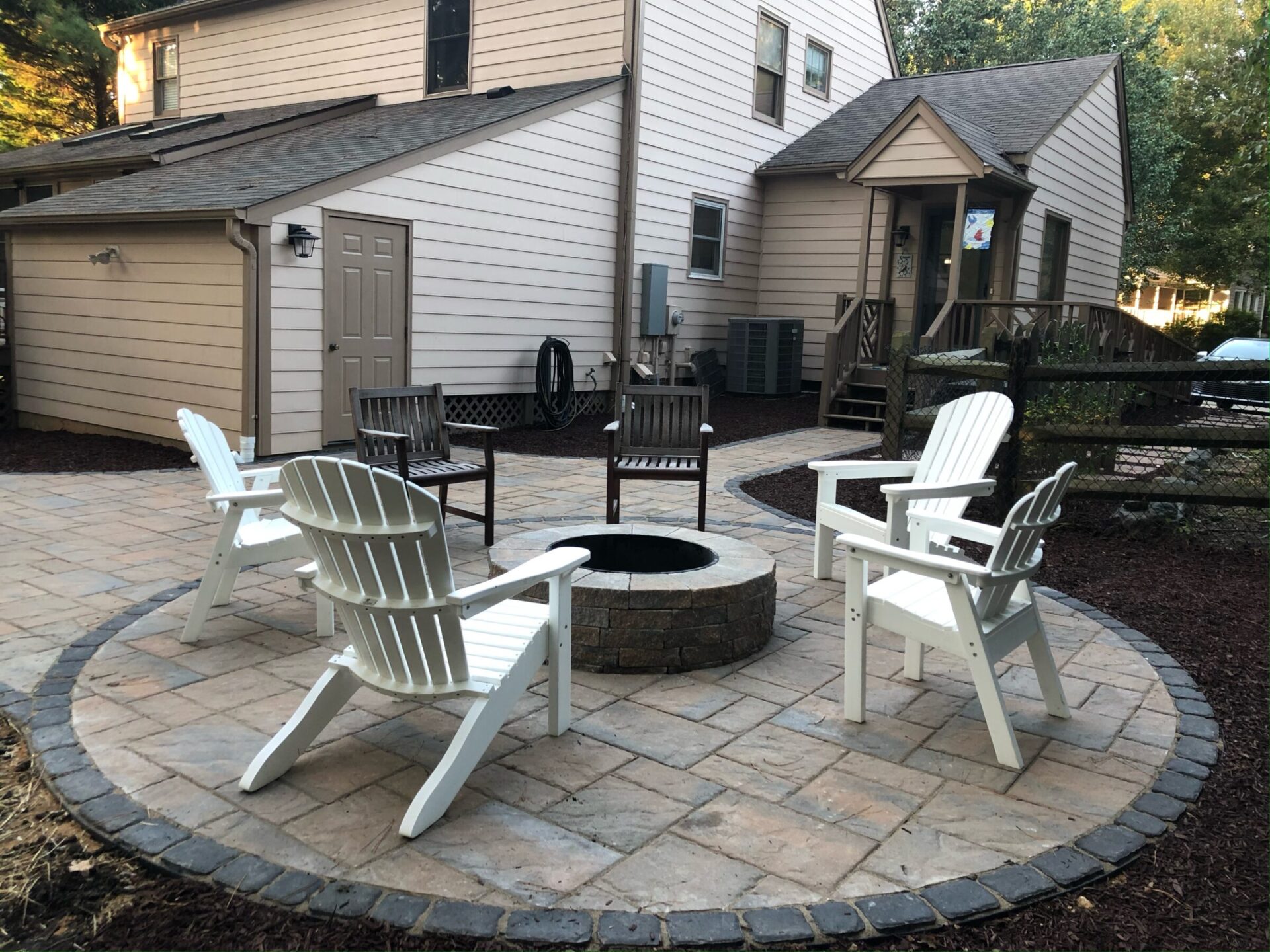 A cozy backyard patio features a circular stone fire pit surrounded by Adirondack chairs and a beige house in the background.