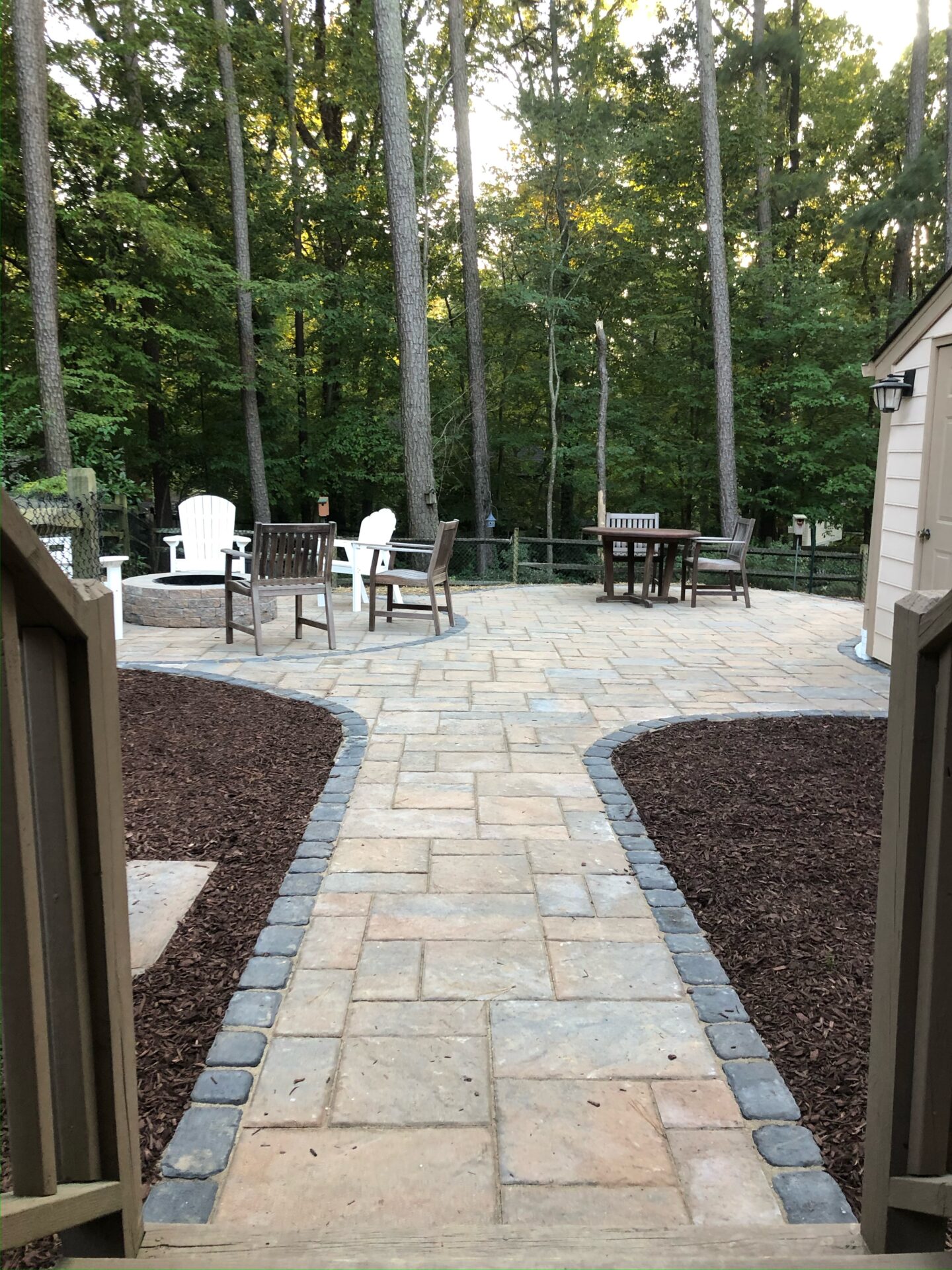 Stone patio with chairs and tables, surrounded by tall trees and mulched paths, creates a peaceful outdoor setting. No recognizable landmarks present.