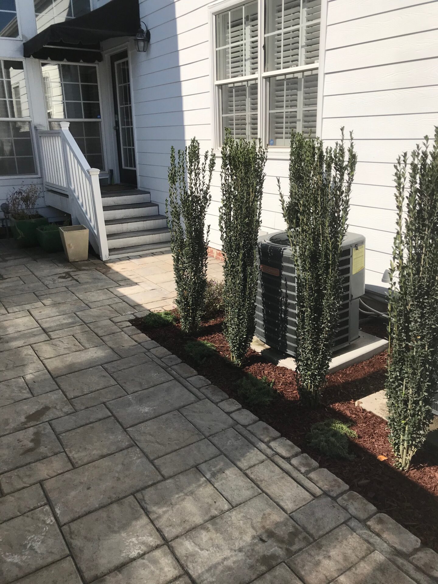 A paved patio with slender shrubs lining an air conditioning unit, adjacent to a white house with steps leading to a door.