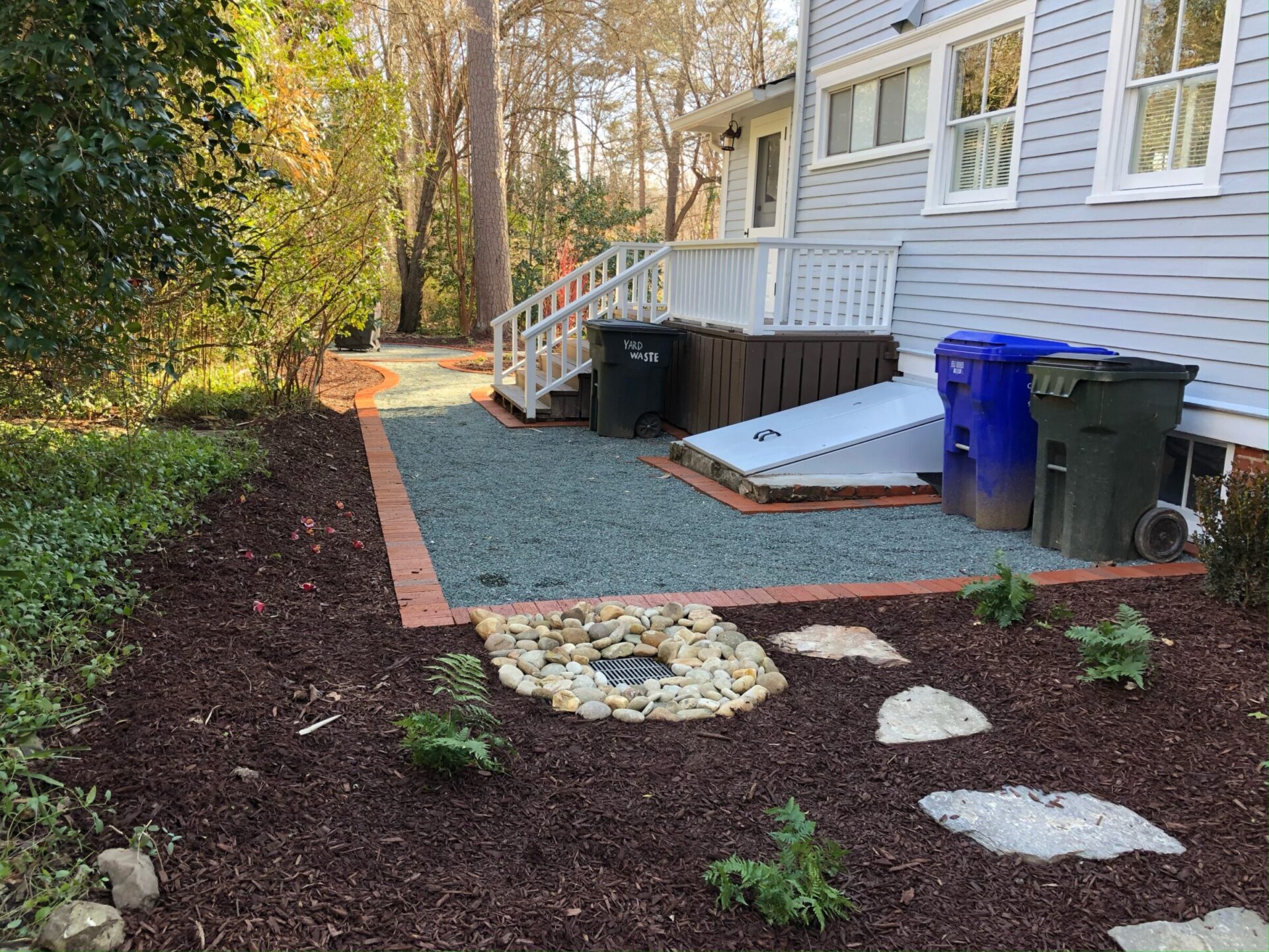 A backyard garden with mulch, rocks, and plants beside a grey house. Trash bins are placed near stairs leading to a back door.