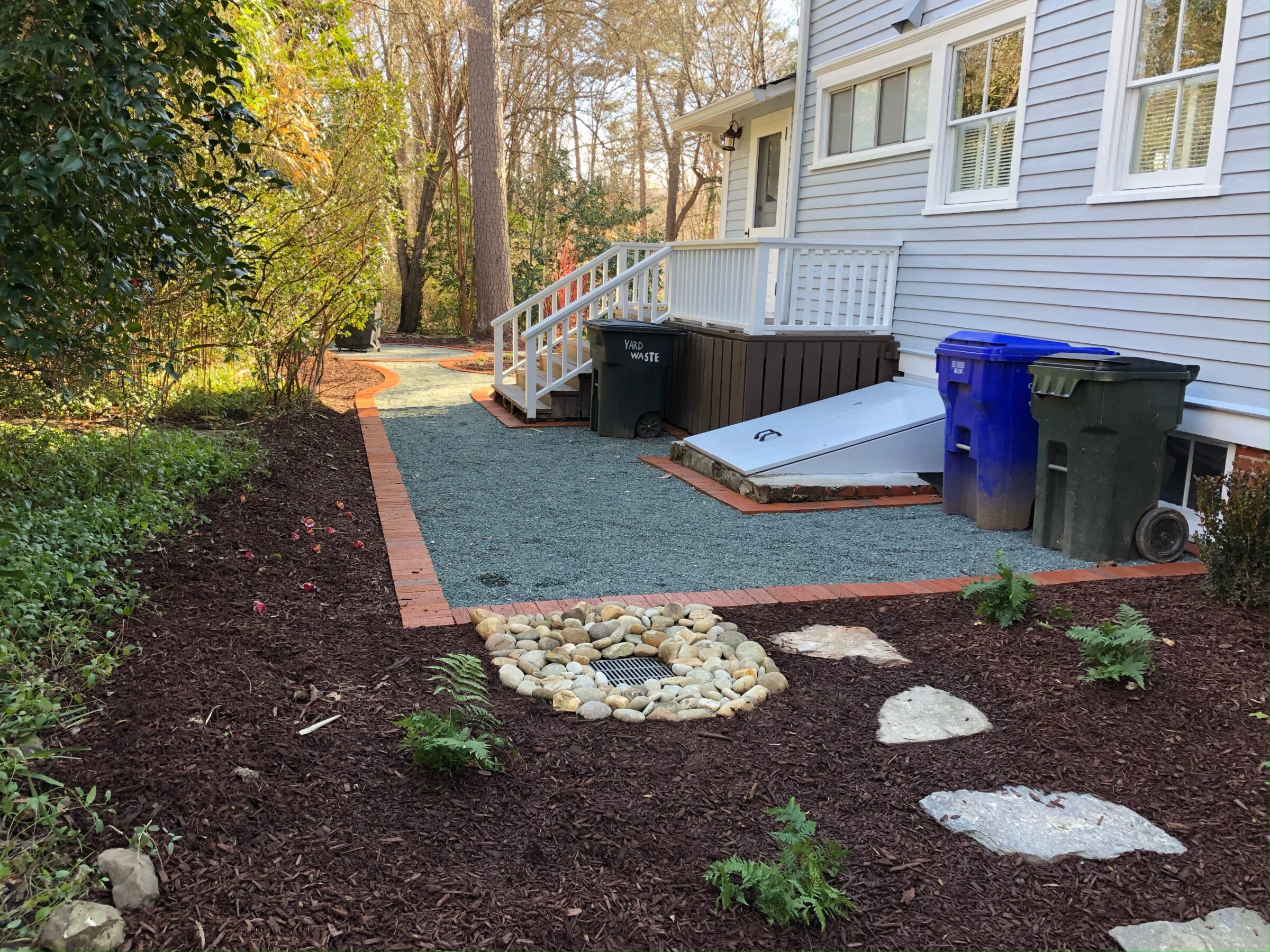 A backyard garden with mulch, rocks, and plants beside a grey house. Trash bins are placed near stairs leading to a back door.