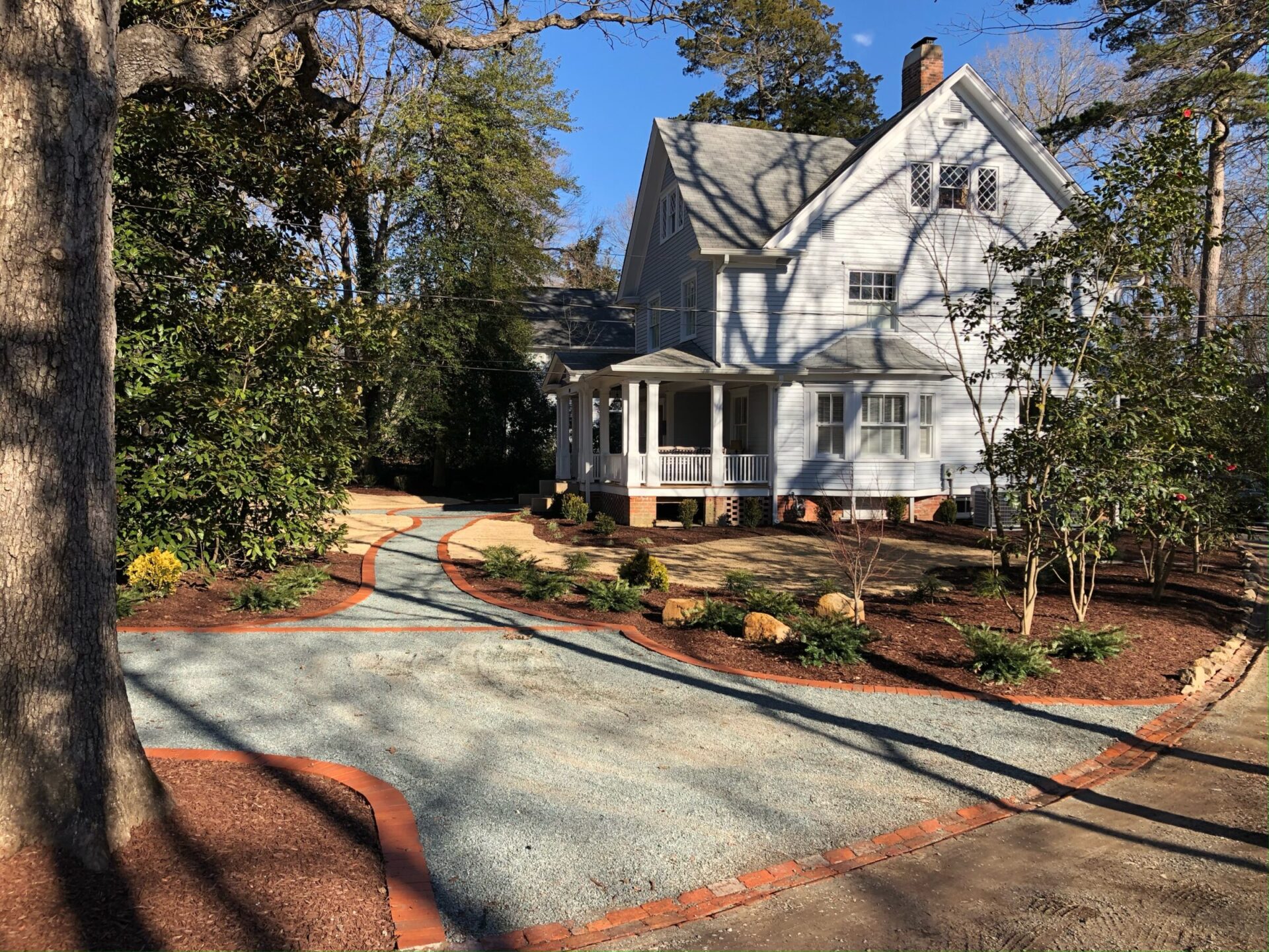 A charming two-story house with a wraparound porch, surrounded by trees and a neatly landscaped yard with a winding gravel pathway.